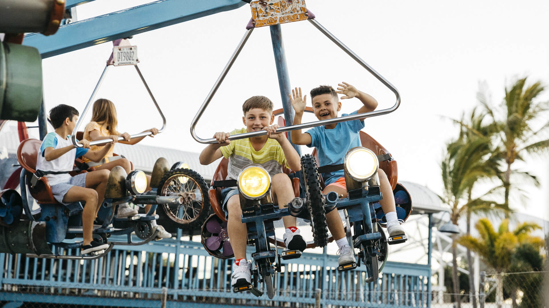 Children ride a spinning amusement park attraction with bicycle-shaped seats, smiling and waving, under a clear sky with palm trees in the background.