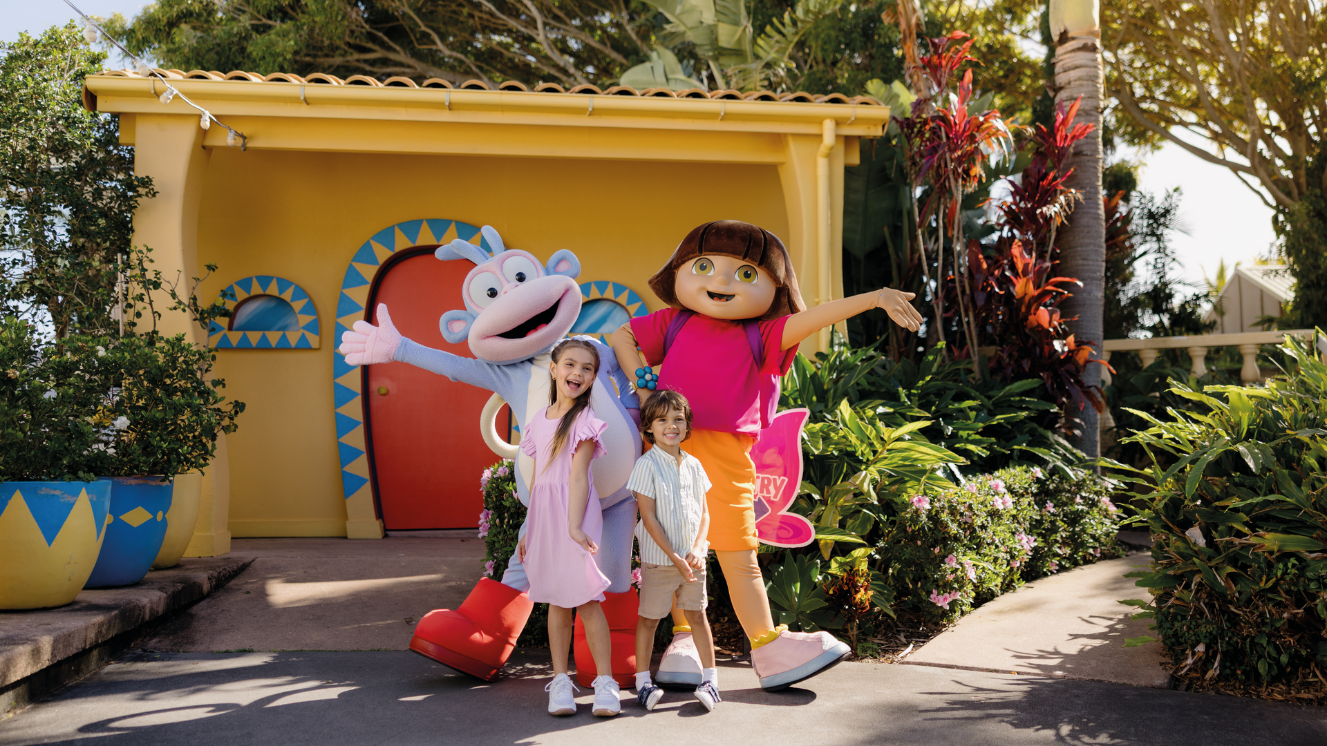 Two children smile and pose outdoors with people dressed in large, colorful costumes of Boots the monkey and Dora the Explorer. They stand in front of a vibrant yellow building surrounded by lush plants and flowers.