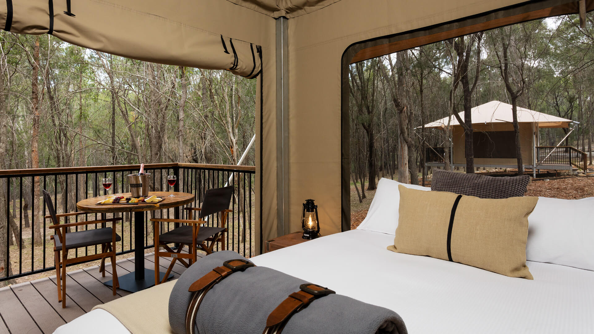A glamping tent features a made bed, a small table with two chairs, wine glasses on a deck, and views of trees and another tent in the background.