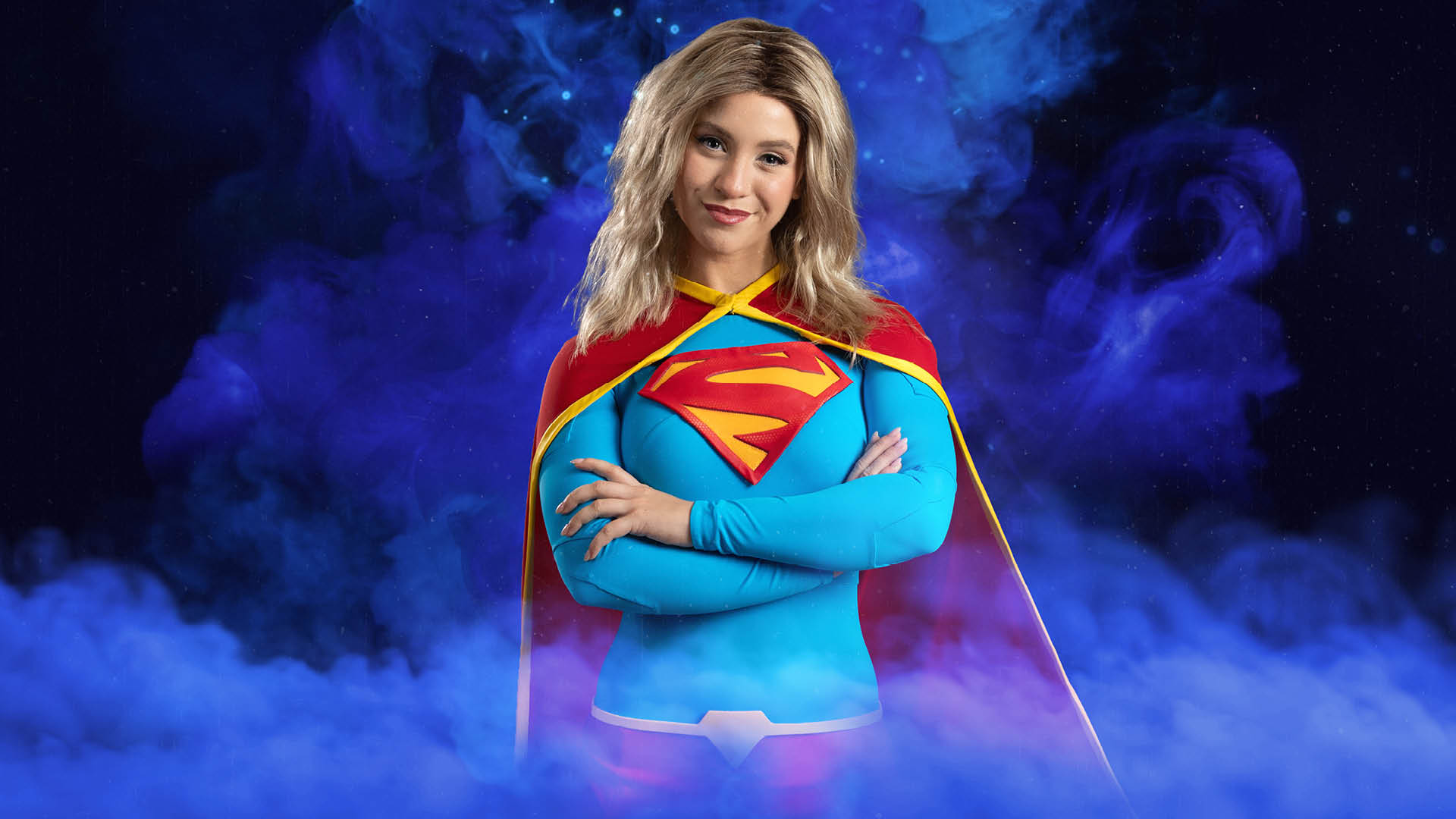 A woman dressed as Supergirl stands with arms crossed, wearing a blue suit with a red and yellow "S" emblem, red cape, and blonde hair, against a smoky blue and black background.