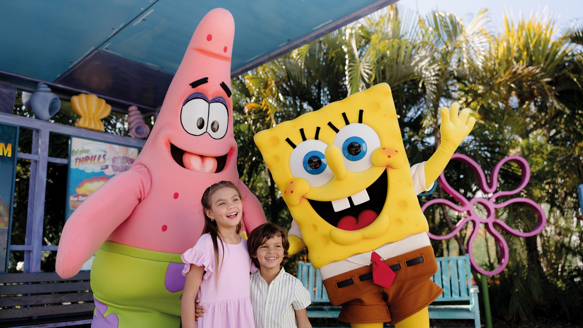 Two children smile and pose with people in large SpongeBob SquarePants and Patrick Star costumes outdoors, surrounded by palm trees and a colorful, theme park-like setting.
