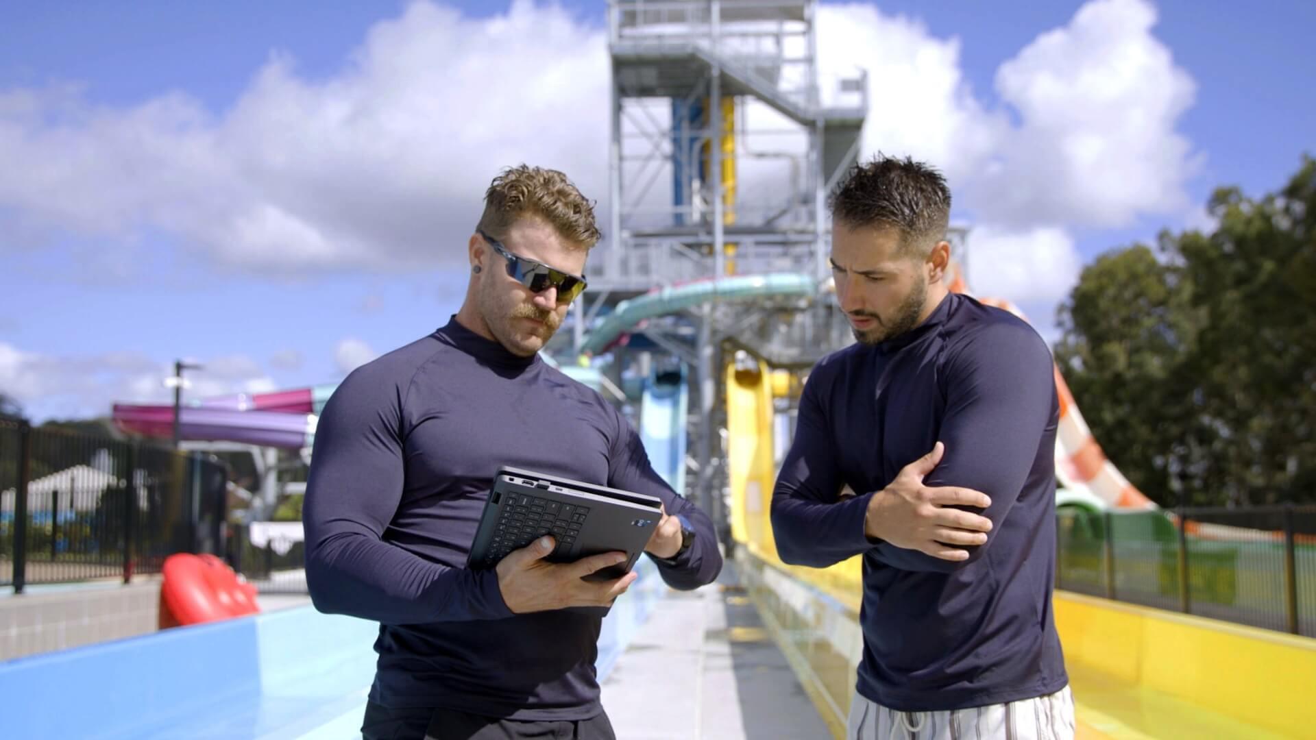 Two men in dark long-sleeve shirts stand at a water park, one holding a tablet and pointing at it while the other crosses his arms and looks on. Water slides and blue sky are visible in the background.