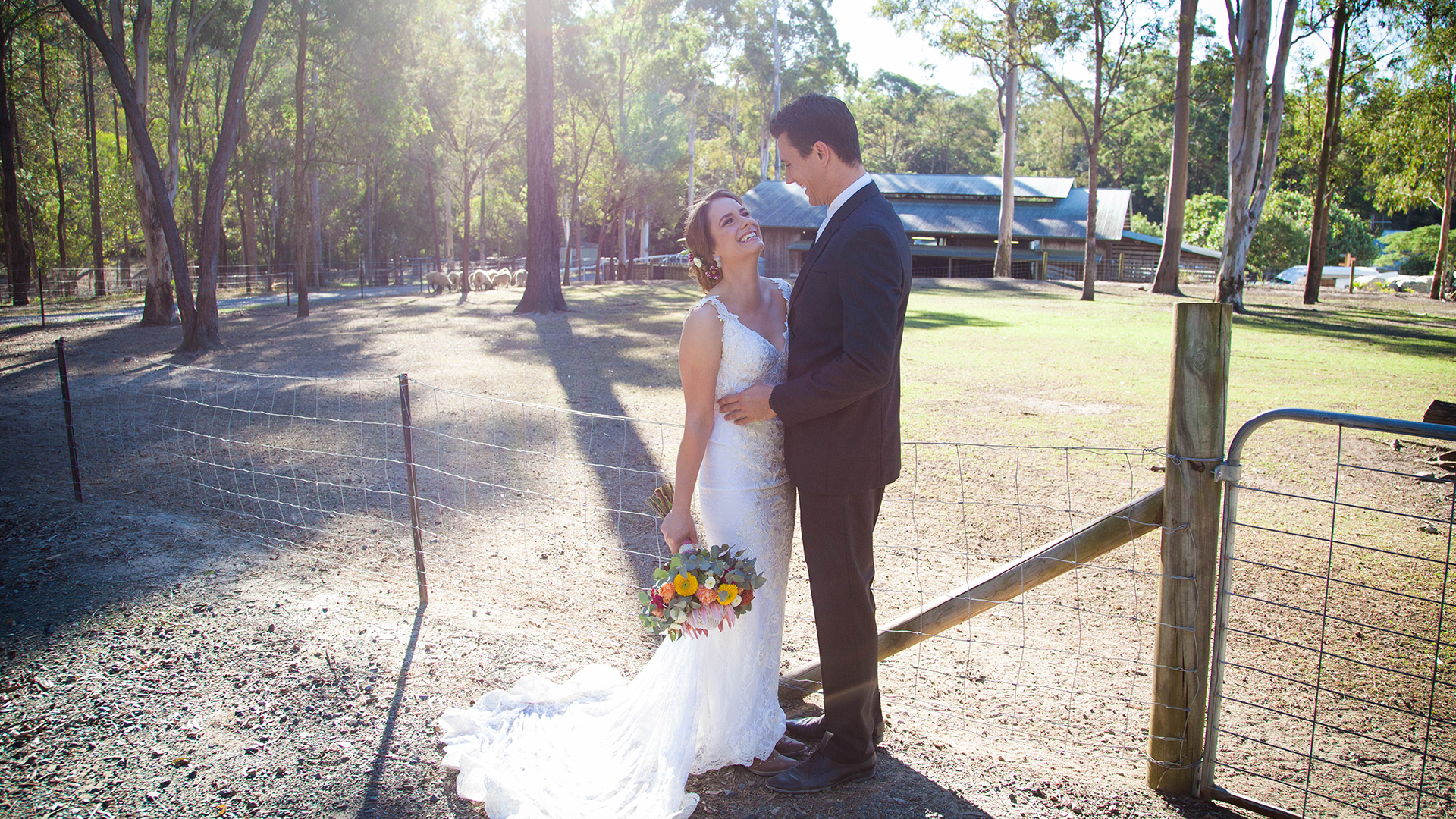 Bride and groom smile at each other. The bride is holding a bouquet of flowers. They are on a rustic farm, standing beside a fence.