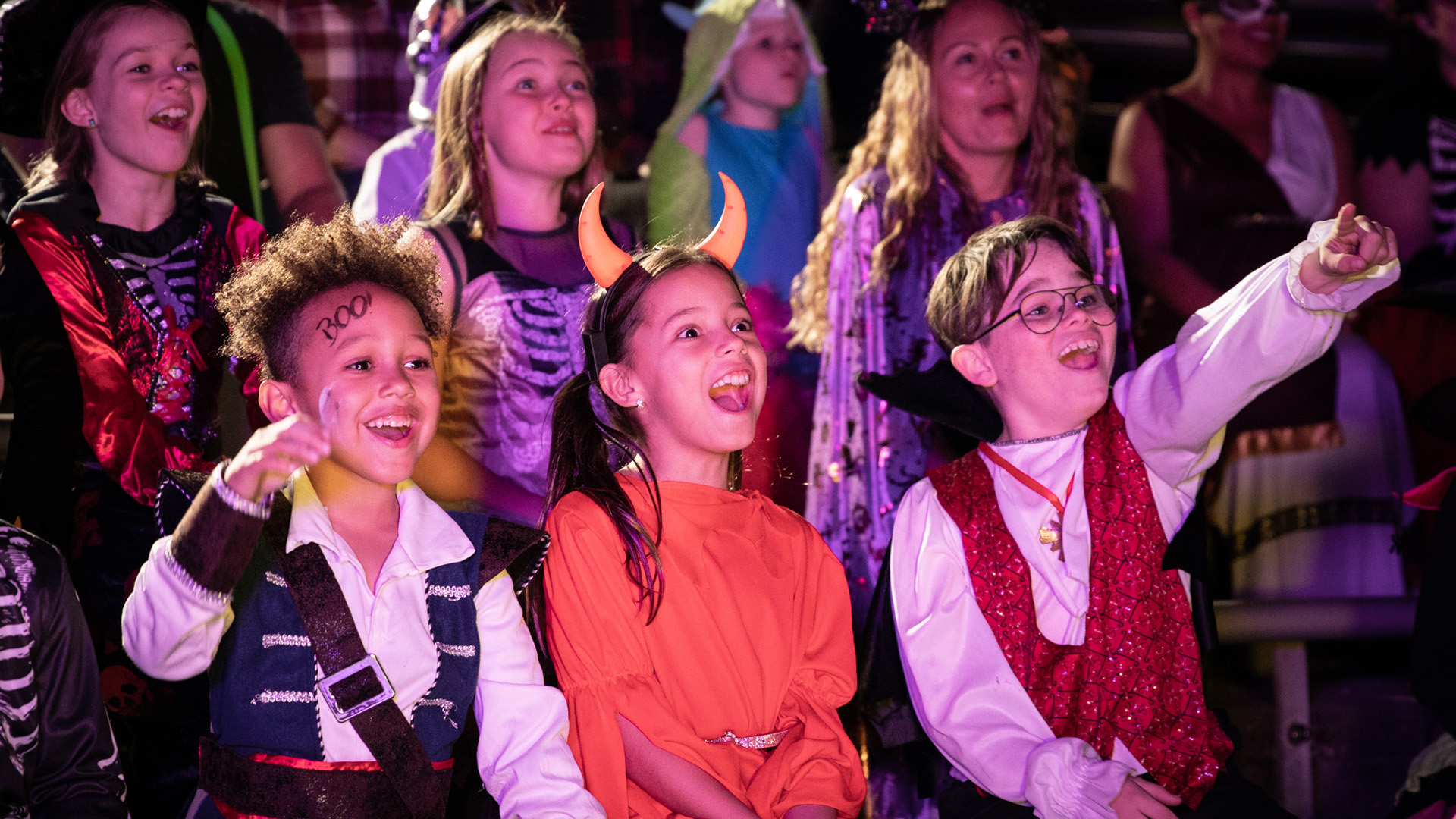 A group of children in colorful Halloween costumes sit together, laughing and cheering excitedly while watching a show or performance. The atmosphere is festive and joyful, with costumes including pirates and devils.