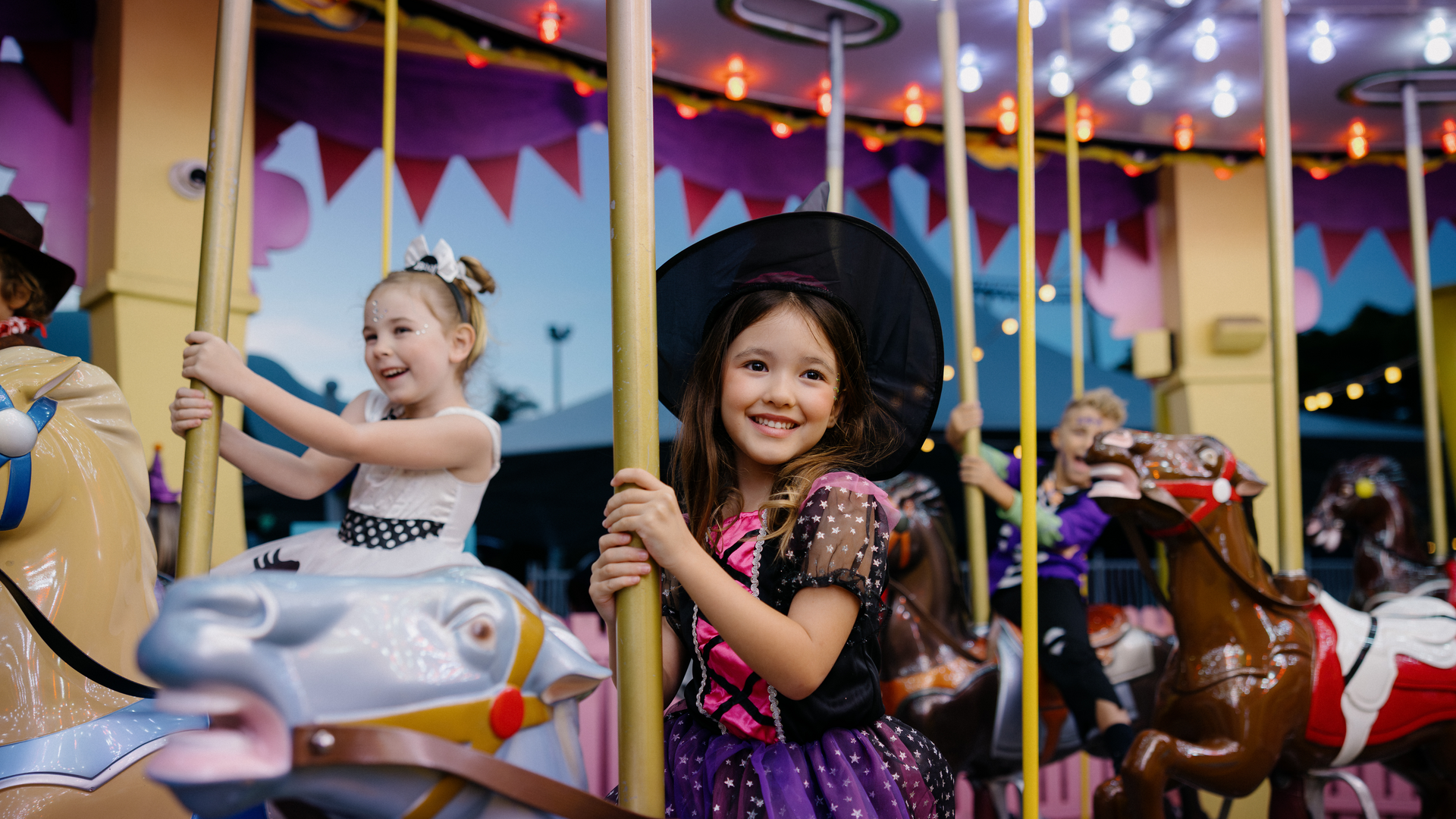 Two young girls in costumes, one dressed as a witch, smile and ride carousel horses at an amusement park. The background has colorful lights and decorations, creating a festive atmosphere.