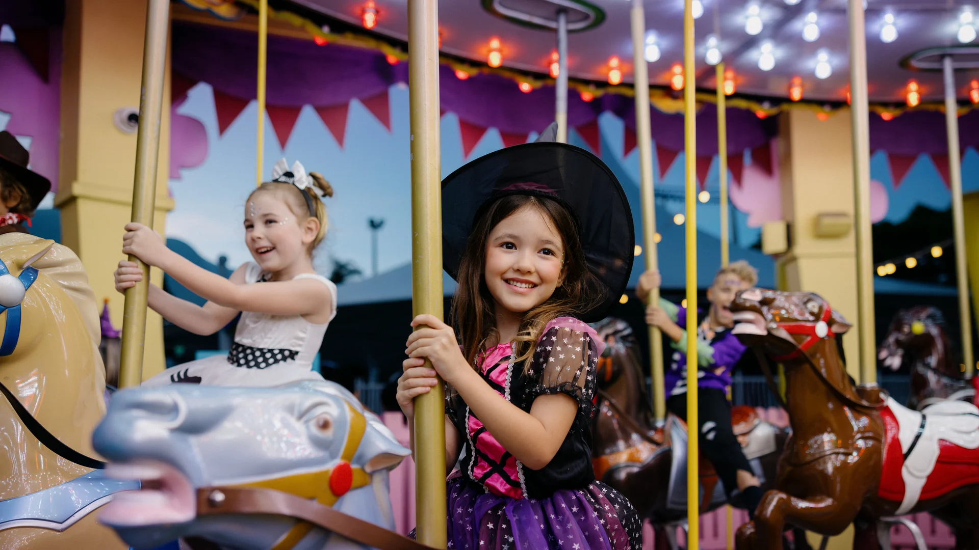 Two young girls in costumes, one dressed as a witch, smile and ride carousel horses at an amusement park. The background has colorful lights and decorations, creating a festive atmosphere.
