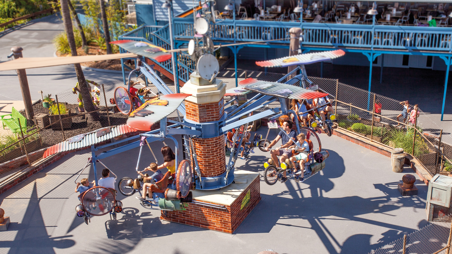 Aerial view of people riding a small outdoor amusement park ride with seats and propeller-themed arms, surrounded by a fence and nearby seating area.