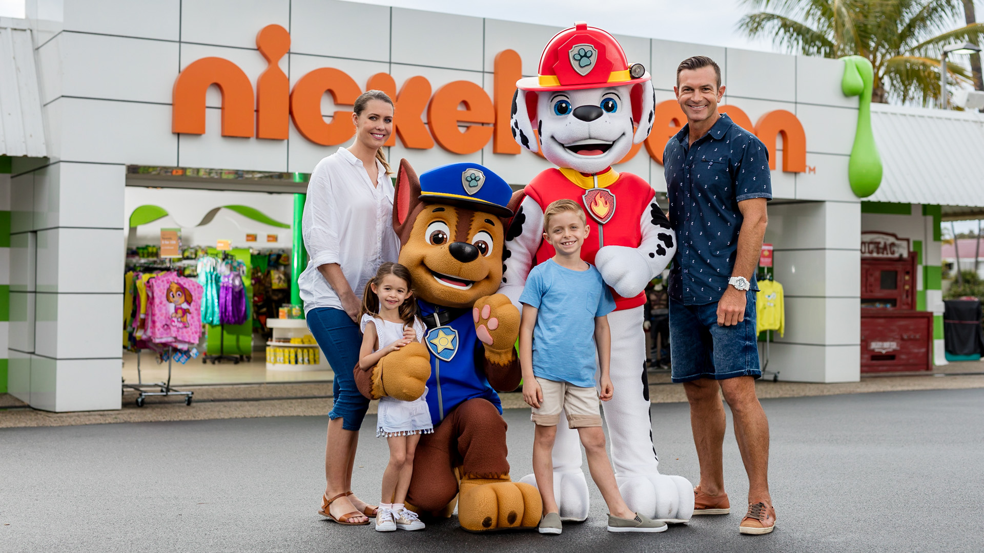 A family of four poses with PAW Patrol characters Chase and Marshall in front of a Nickelodeon store at an outdoor location.