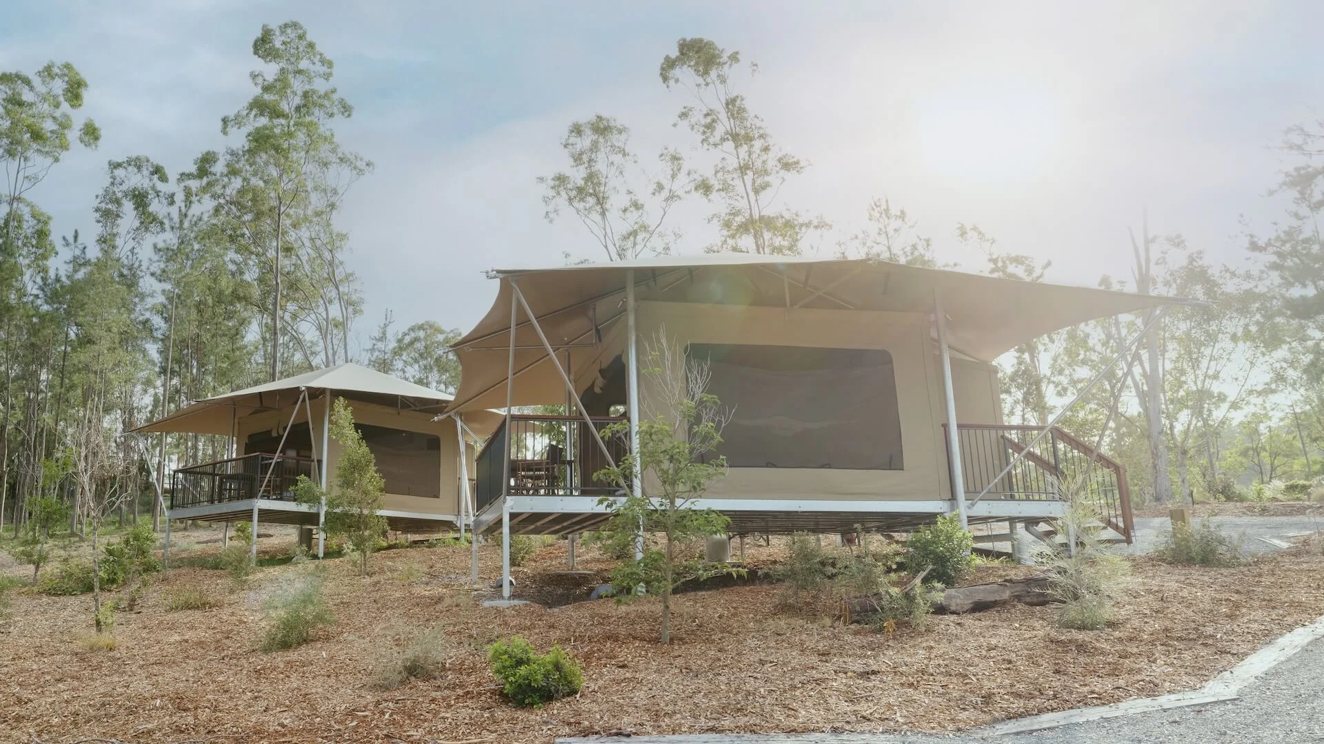 Two canvas glamping tents on raised platforms are set among trees, with sunlight filtering through a partly cloudy sky.