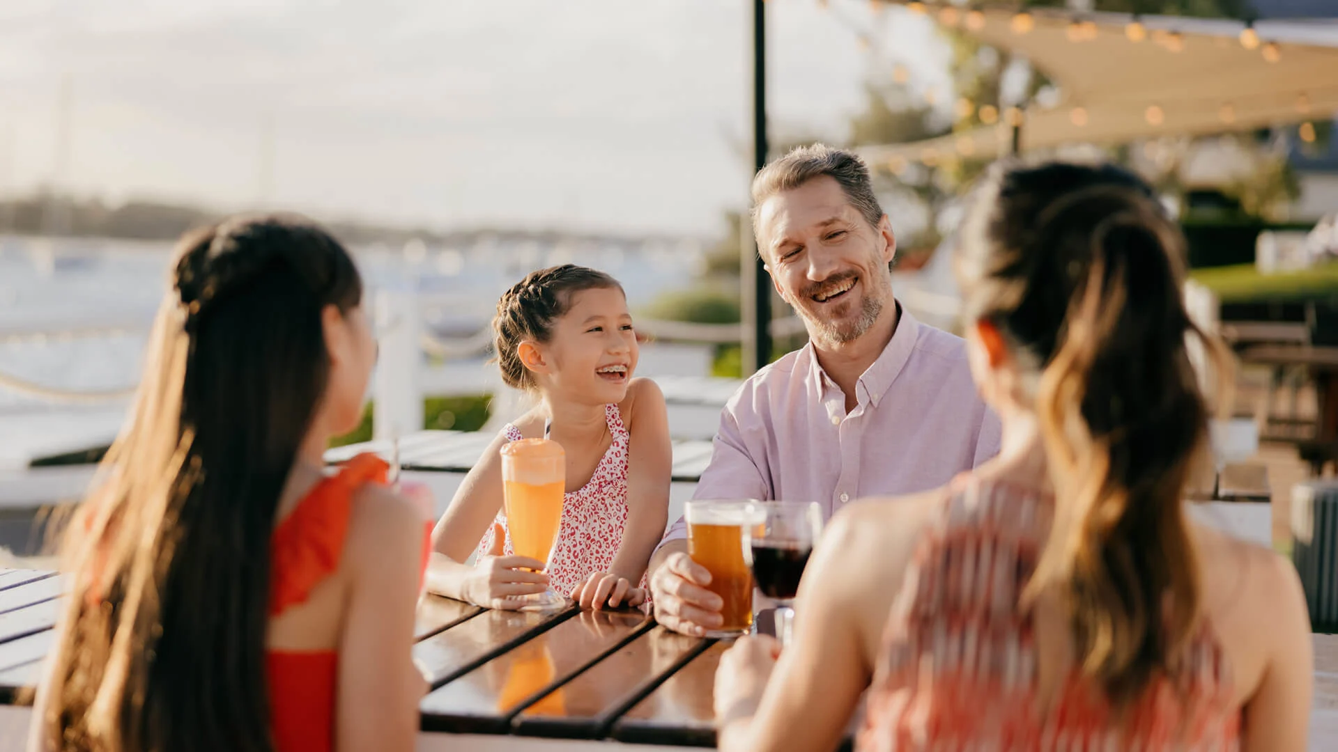 A family of four sits at an outdoor table at the Boat Shed, Sea World Resort, enjoying drinks and smiling together in a sunny setting.