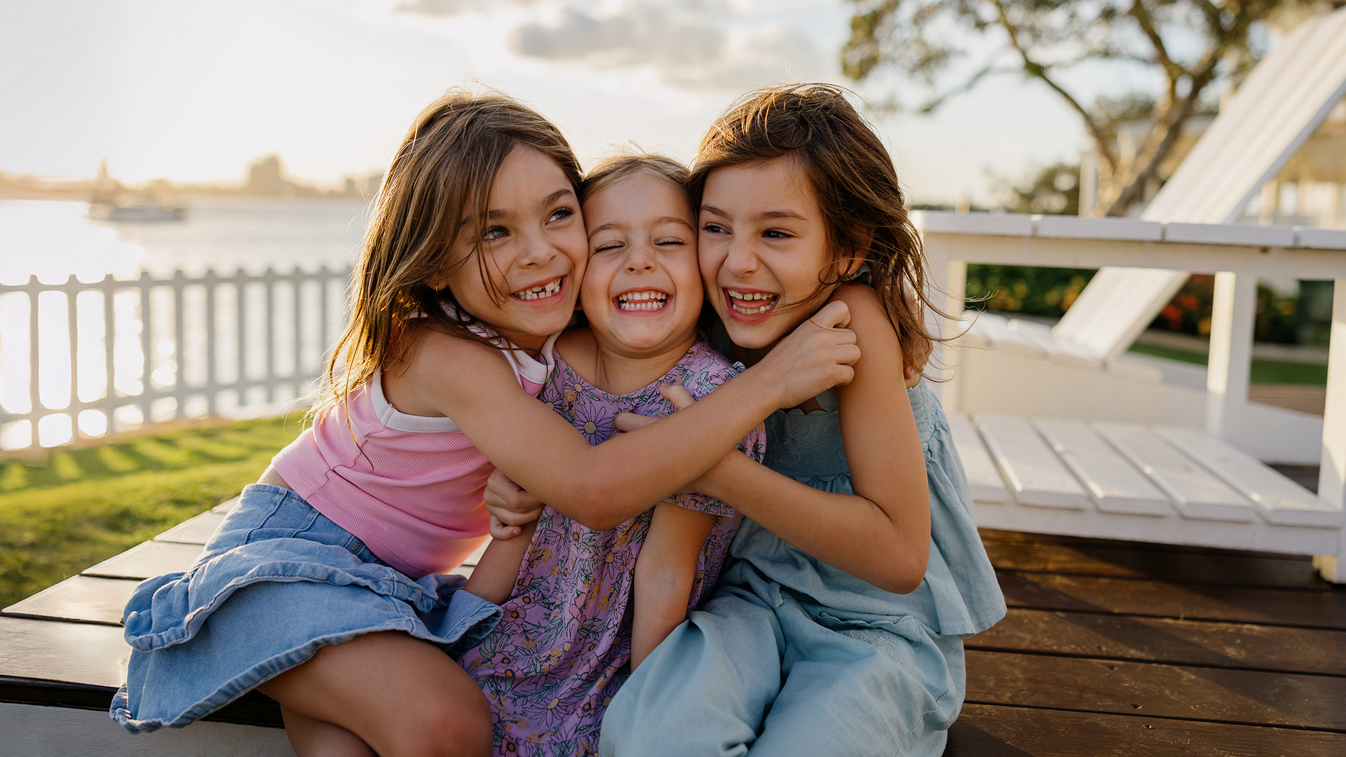 Three young girls sit outdoors on a wooden deck near a white fence, smiling and hugging each other while enjoying a sunny day by the water.