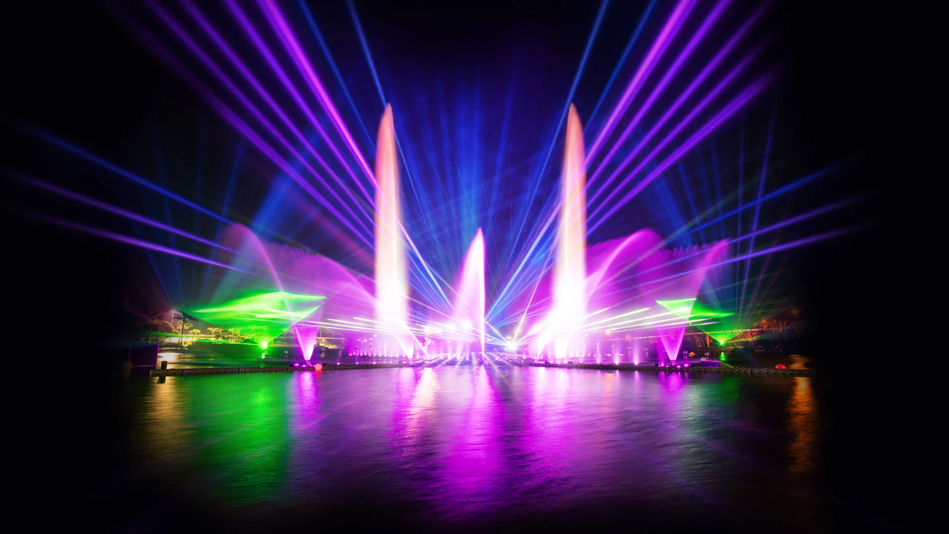 A vibrant nighttime light show over water features colorful laser beams, illuminated fountains, and reflections in green, purple, blue, and pink, creating a dynamic and dazzling display against a dark background.