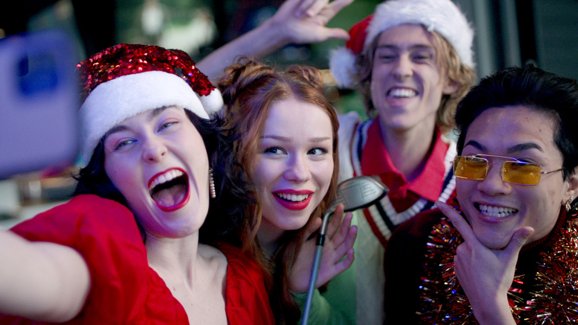 Four young adults in festive outfits and Santa hats smile and pose for a selfie at a holiday party, looking cheerful and excited. One person holds a golf club, and others wear tinsel and holiday accessories.