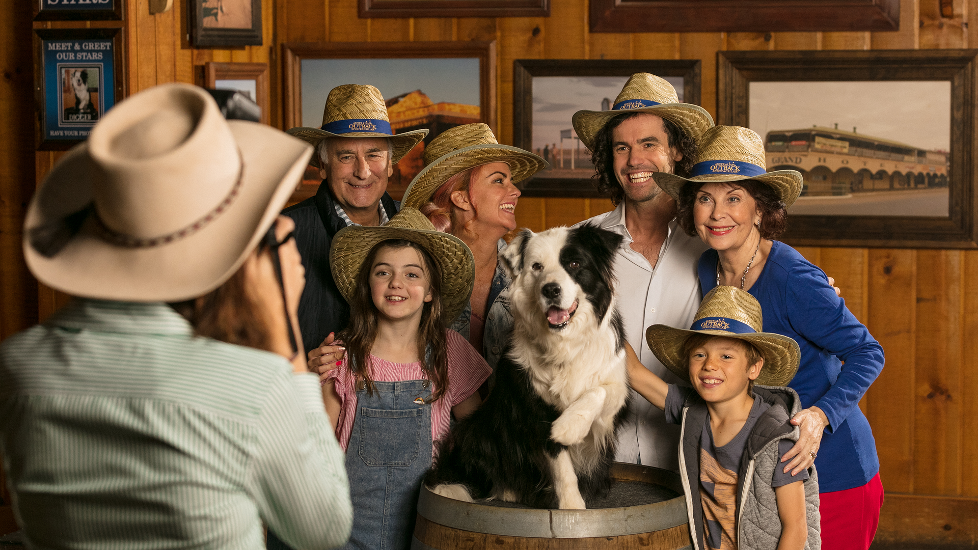 A group of six smiling people, including children and adults, pose indoors with a black-and-white dog sitting in a barrel. They all wear straw hats and stand in front of wooden walls decorated with framed pictures.