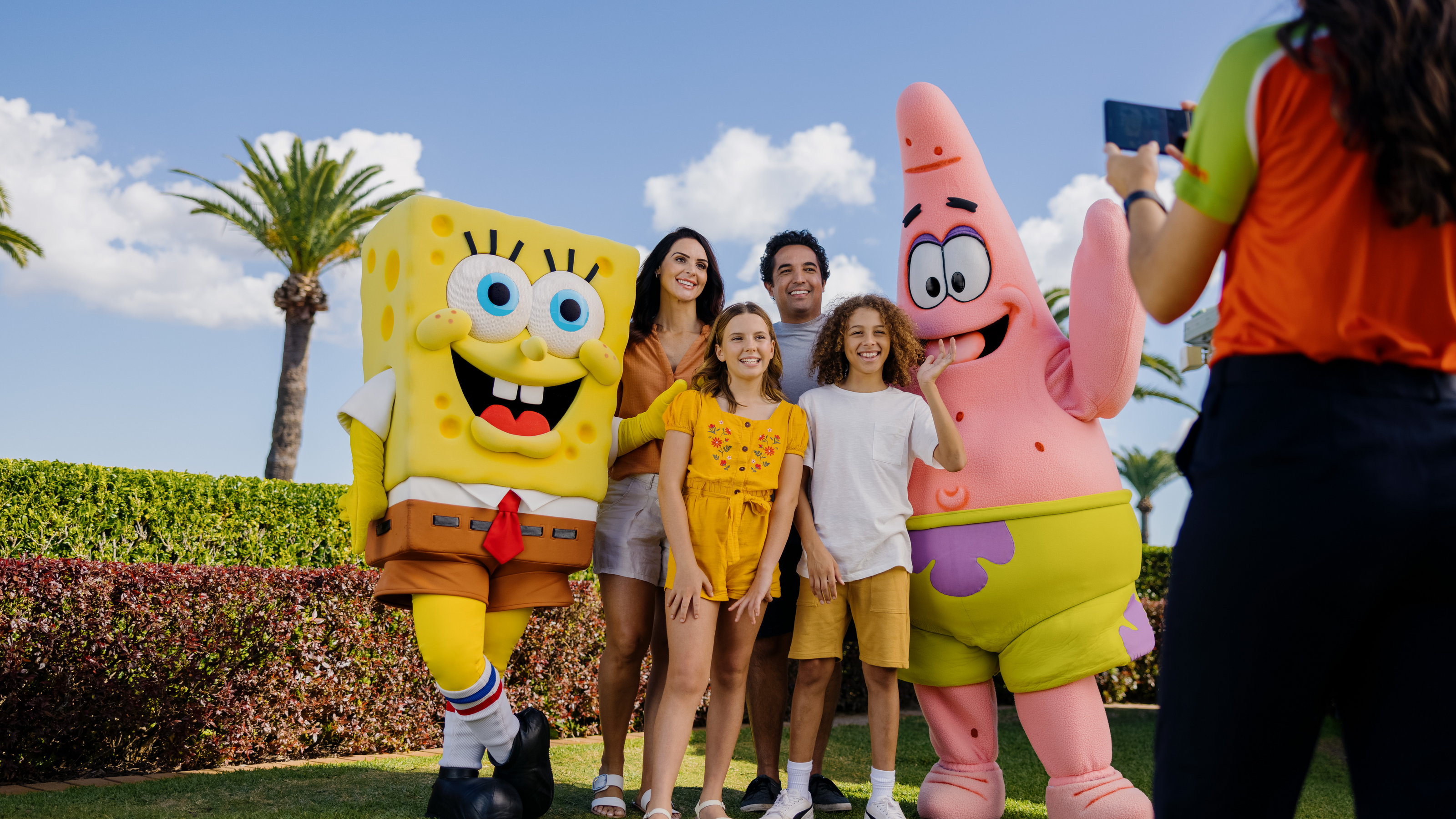 Family poses with SpongeBob and Patrick outdoors as a person takes their photo. Palm trees and hedges are visible in the background.