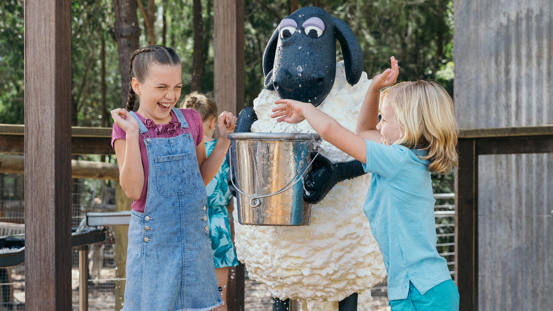 Two children laugh and splash water next to a person in a sheep costume holding a bucket, outdoors by a wooden fence.