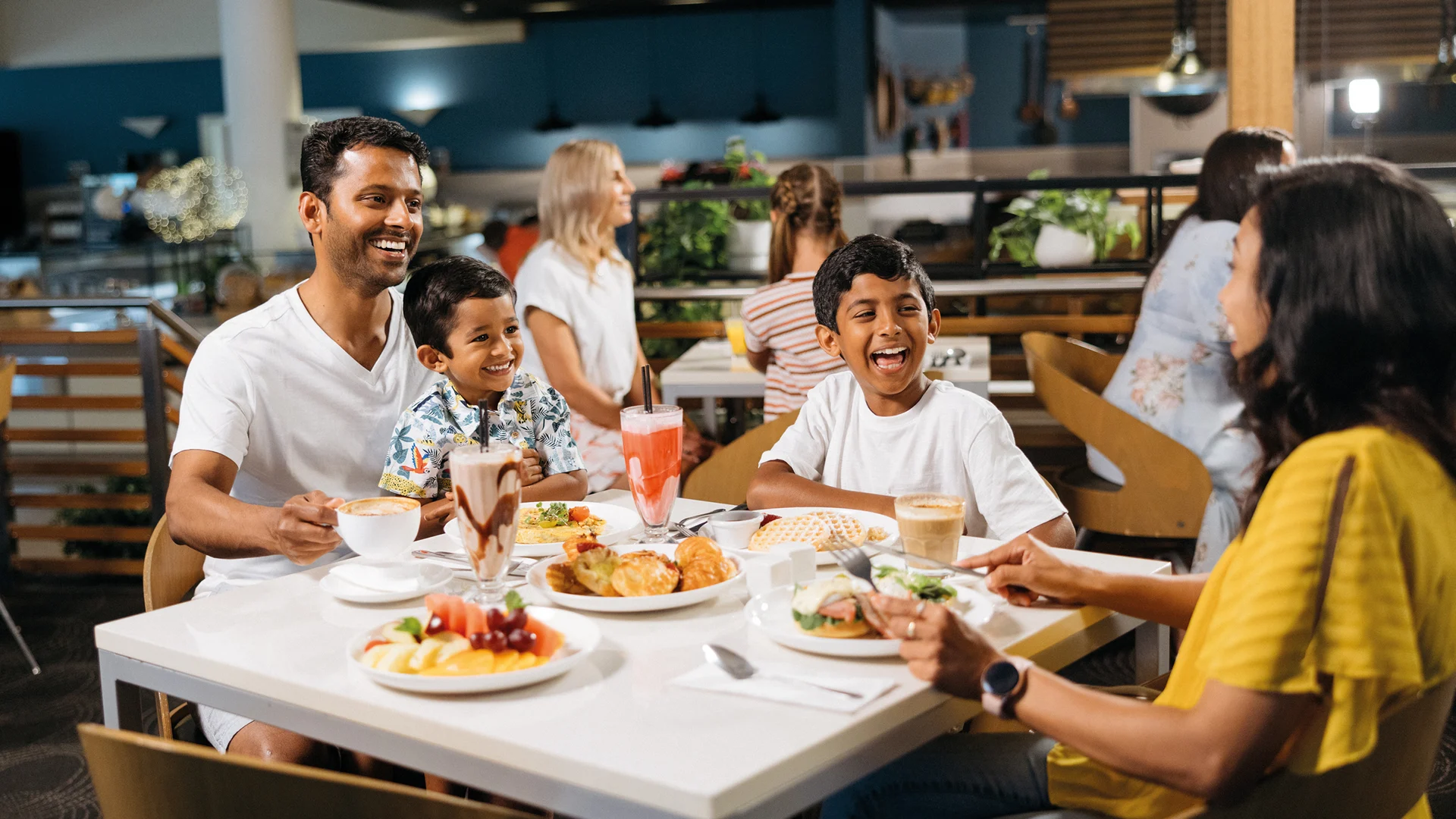 A family of four, including two children and two adults, sits at a restaurant table, smiling and enjoying a meal with various dishes and drinks. Other diners are visible in the background.