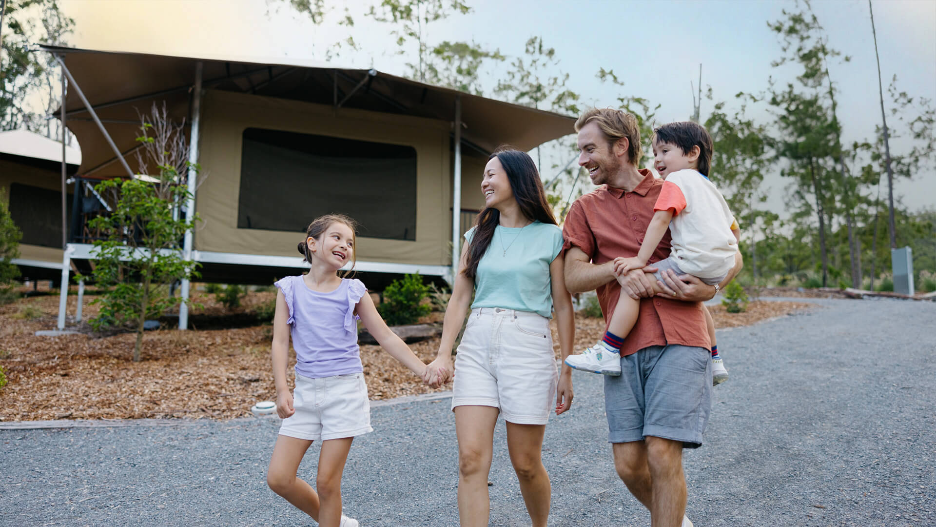 A family of four walks together on a gravel path near a tent cabin in a wooded campsite, smiling and holding hands.