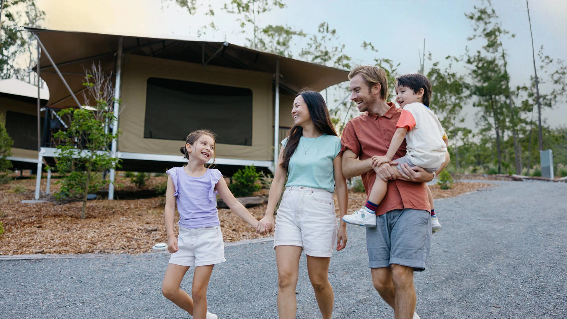 A family of four walks together on a gravel path near a tent cabin in a wooded campsite, smiling and holding hands.