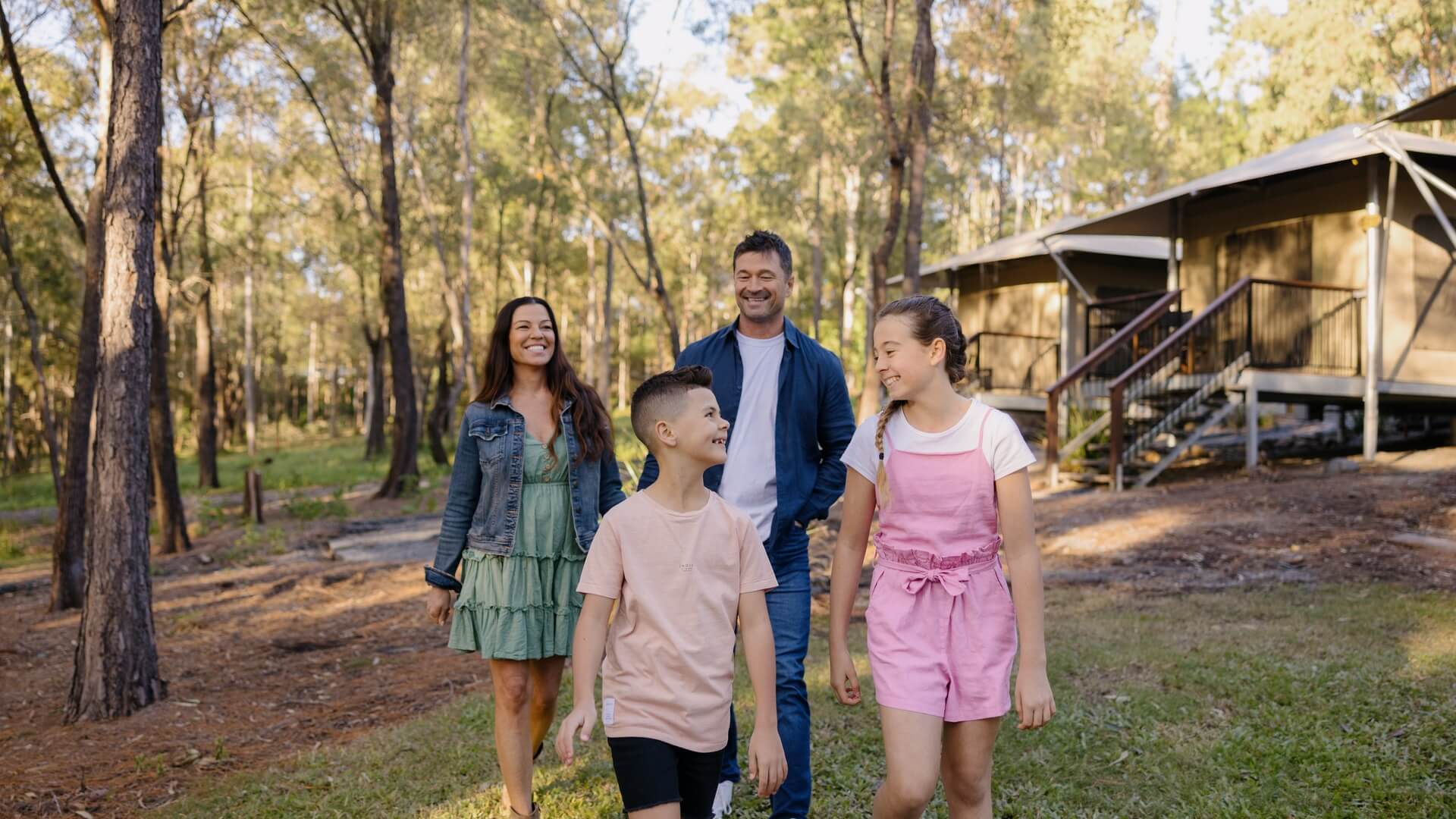 Four people, two adults and two children, walk together outside near a cabin in a wooded area on a sunny day.
