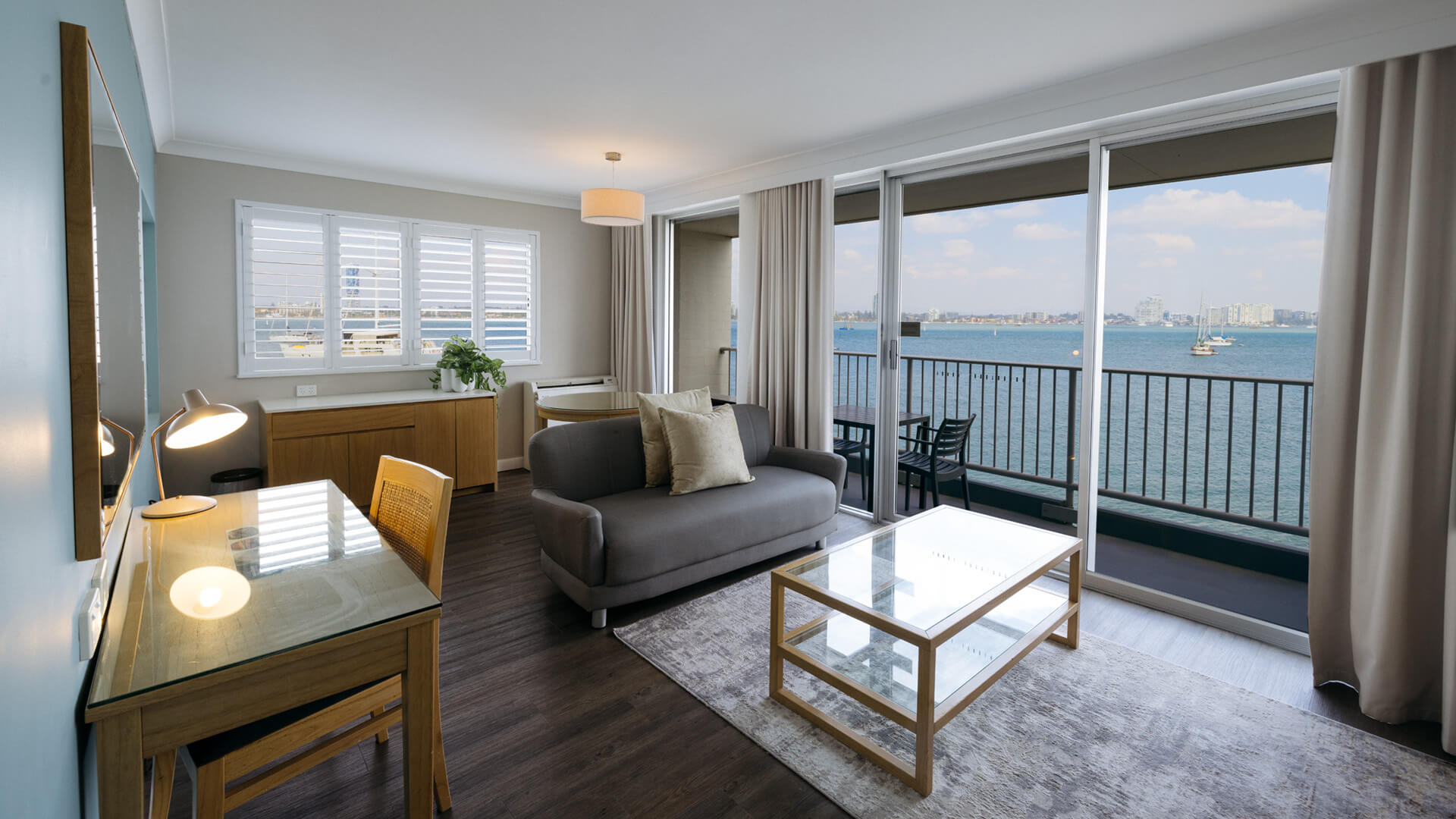 A living room with a grey sofa, glass coffee table, desk, and a large window offering a view of a blue ocean and city skyline.