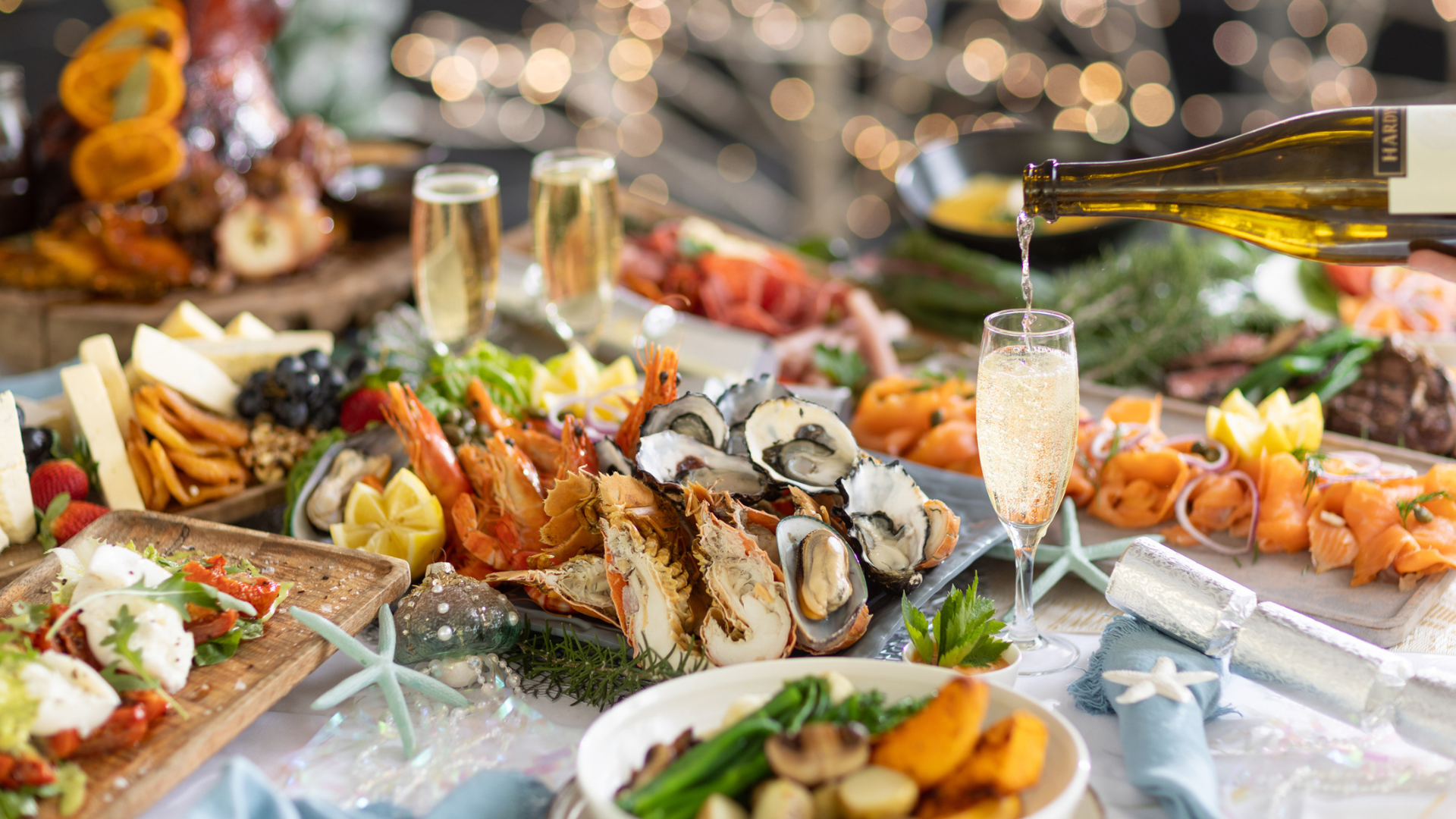 A festive table is set with seafood, cheeses, meats, and champagne being poured into glasses. The background has sparkling lights, creating a celebratory atmosphere.