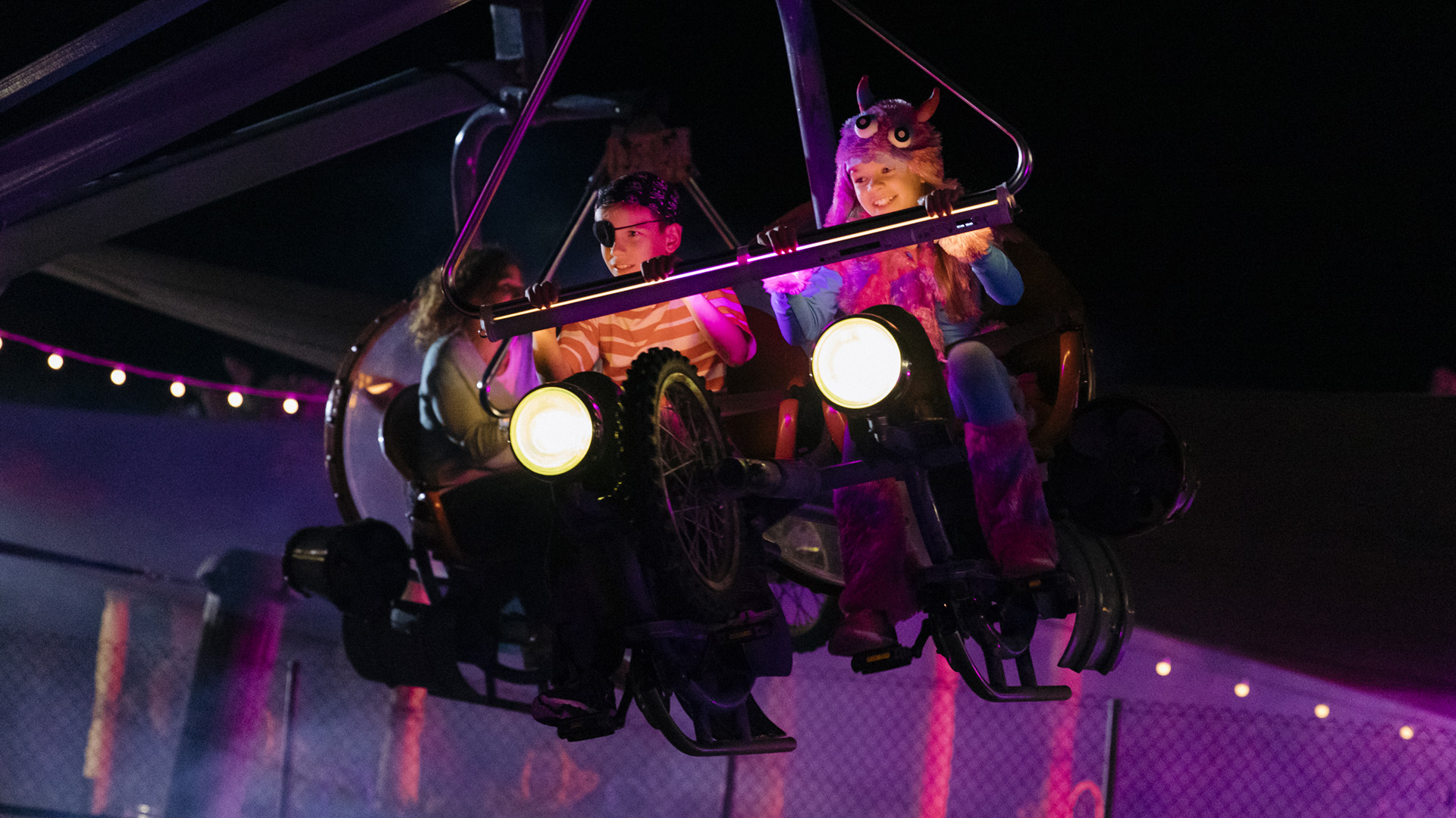 Two people in colorful costumes ride a motorcycle-themed amusement park ride at night, illuminated by bright lights, with a dark background and festive string lights behind them.