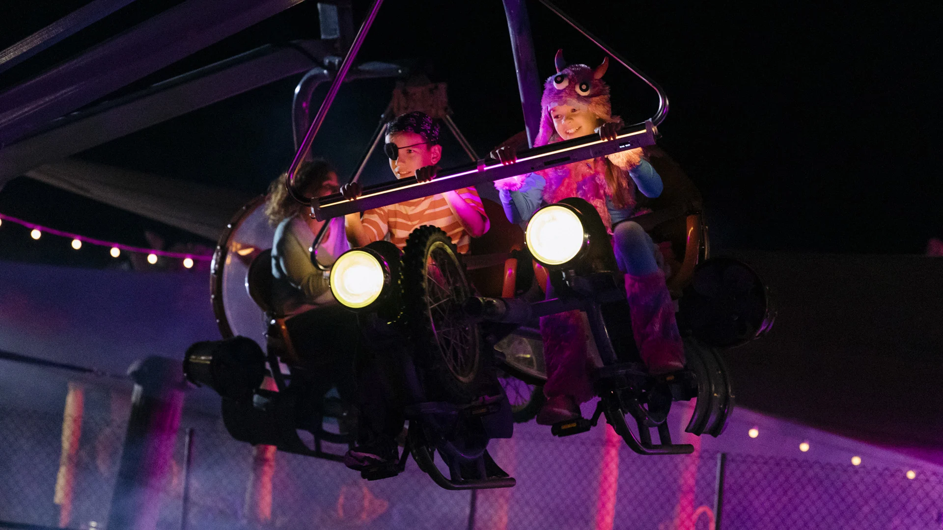 Two people in colorful costumes ride a motorcycle-themed amusement park ride at night, illuminated by bright lights, with a dark background and festive string lights behind them.