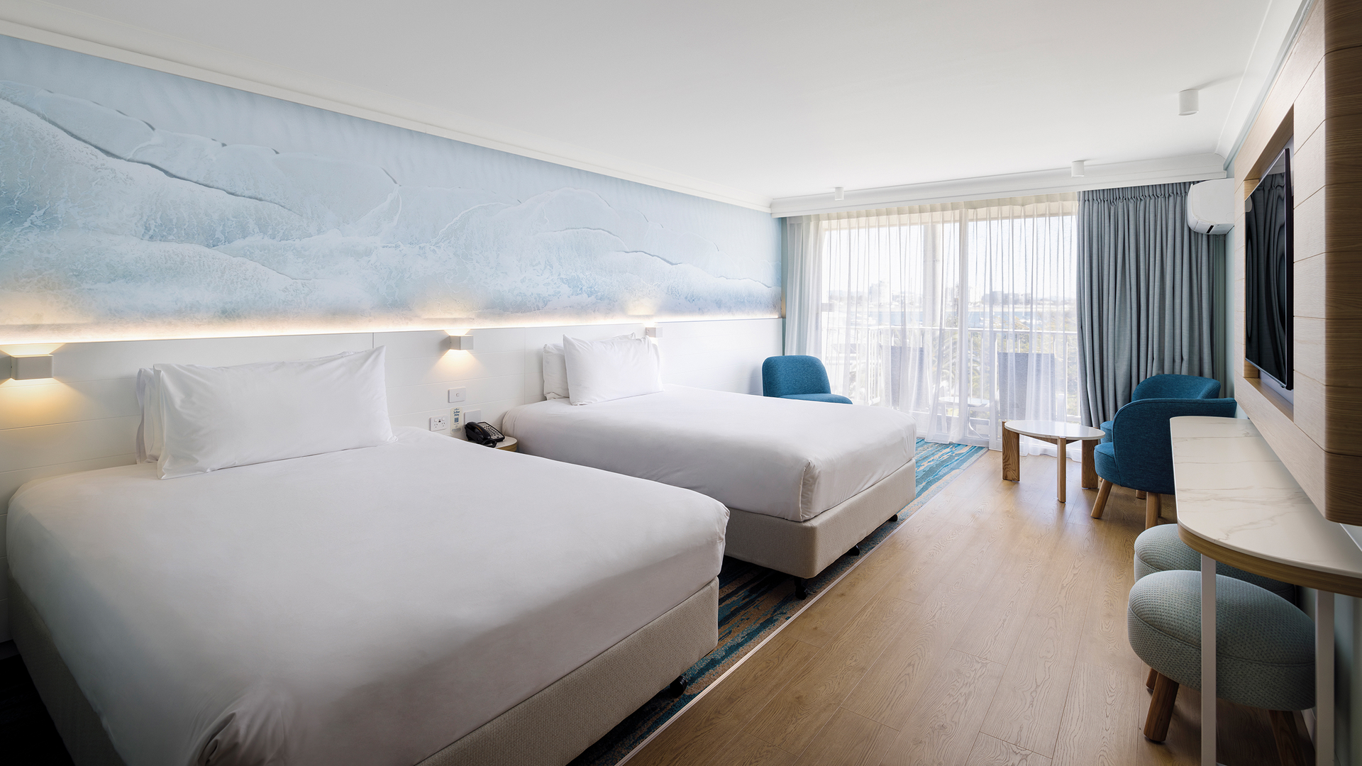 A modern hotel room with two double beds, white linens, a large wall mural, wooden floors, a blue armchair, a round table, a flat-screen TV, and large windows with sheer curtains letting in natural light.