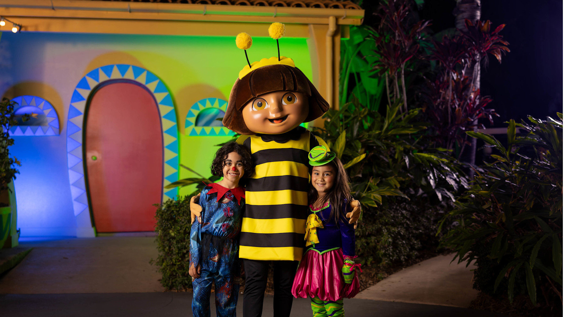 Two children dressed in costumes pose with a person in a bee mascot costume in front of a colorful building.