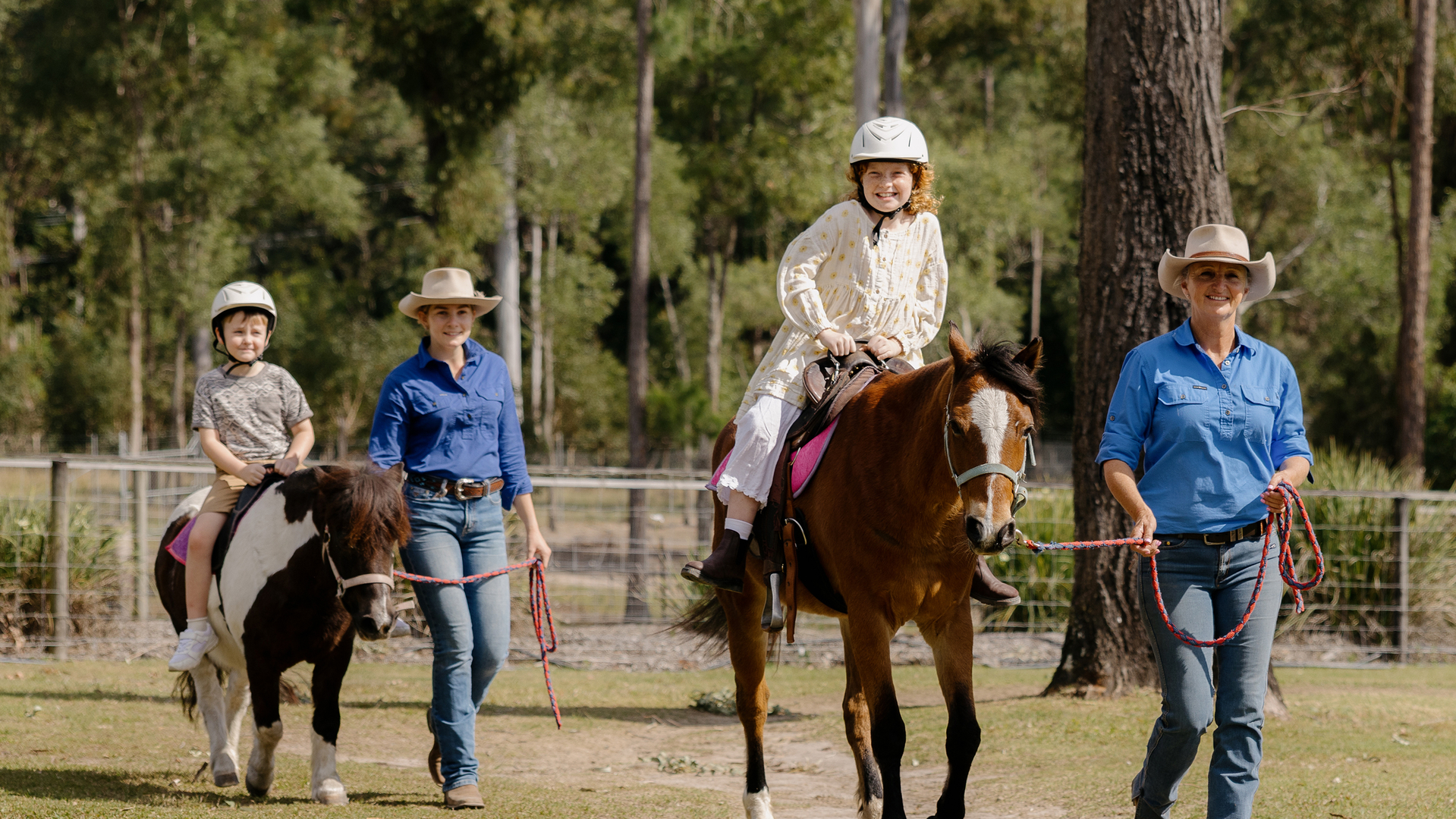 Two children wearing helmets ride horses, each led by an adult in blue shirts and hats, walking on grass in an outdoor setting with trees in the background.