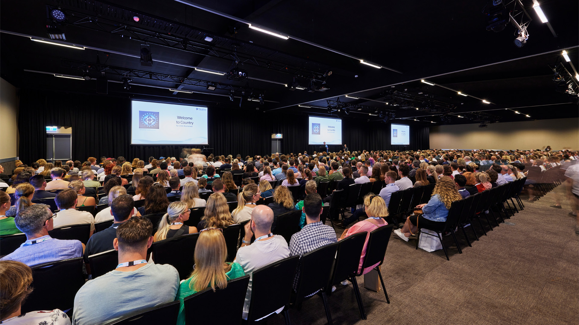 A large audience sits in a conference hall facing multiple presentation screens.