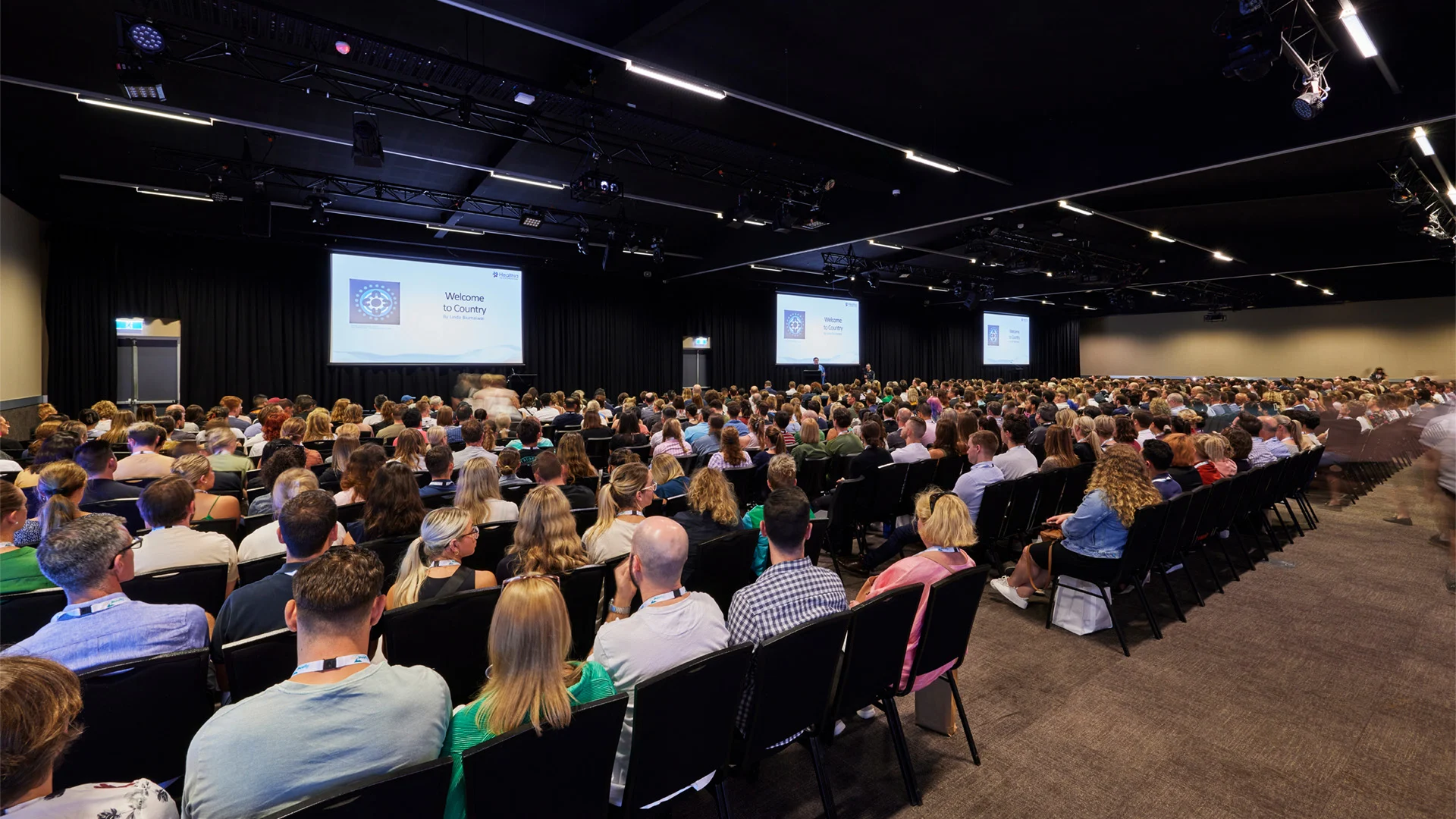 A large audience sits in a conference hall facing multiple presentation screens.