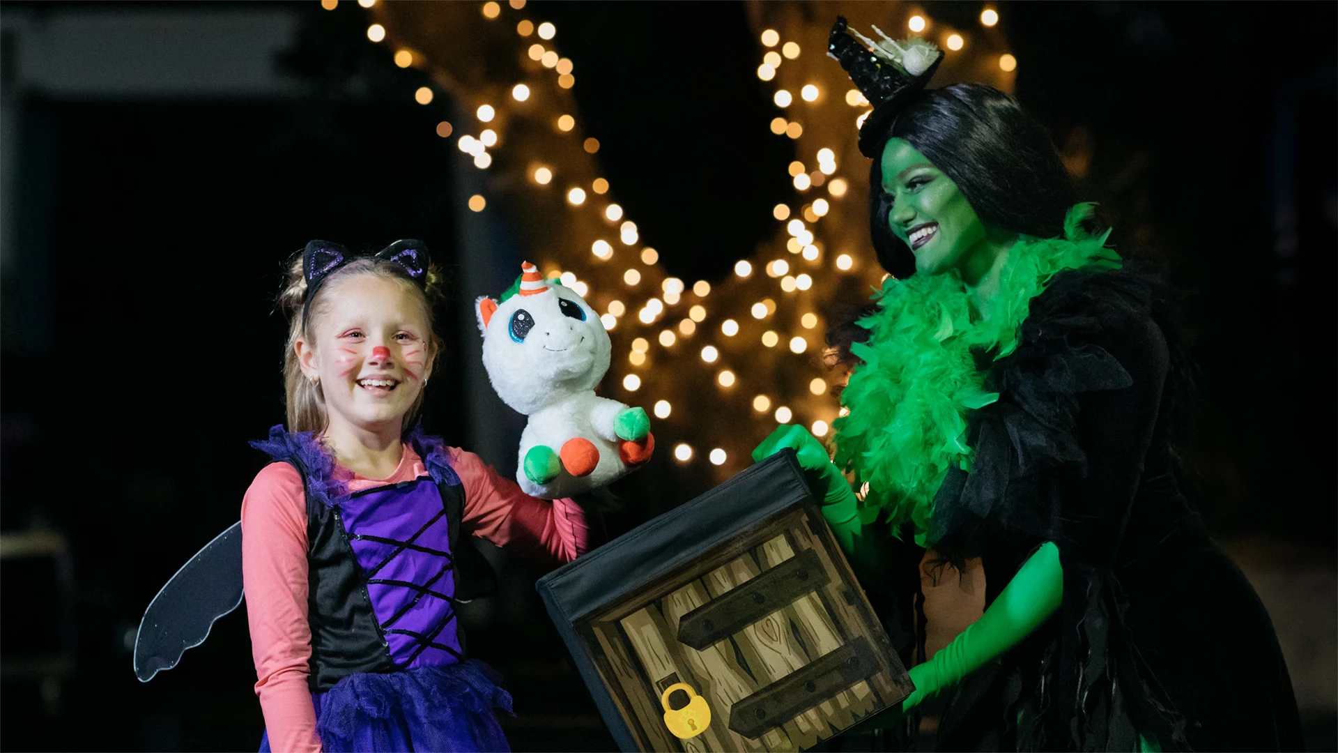 A smiling child in a cat costume holds a stuffed unicorn while standing next to an adult dressed as a green witch, both in front of a tree decorated with string lights.