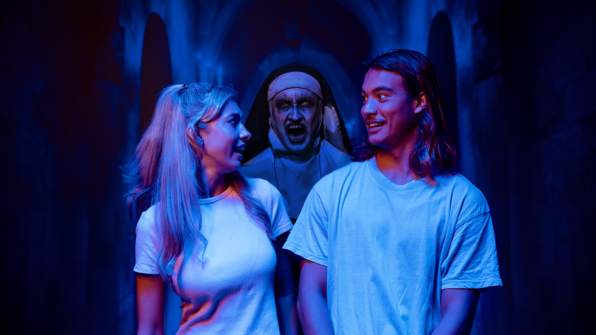 A young man and woman in white shirts smile at each other, unaware of a frightening, ghostly nun looming behind them in a dark, eerie hallway with blue lighting.