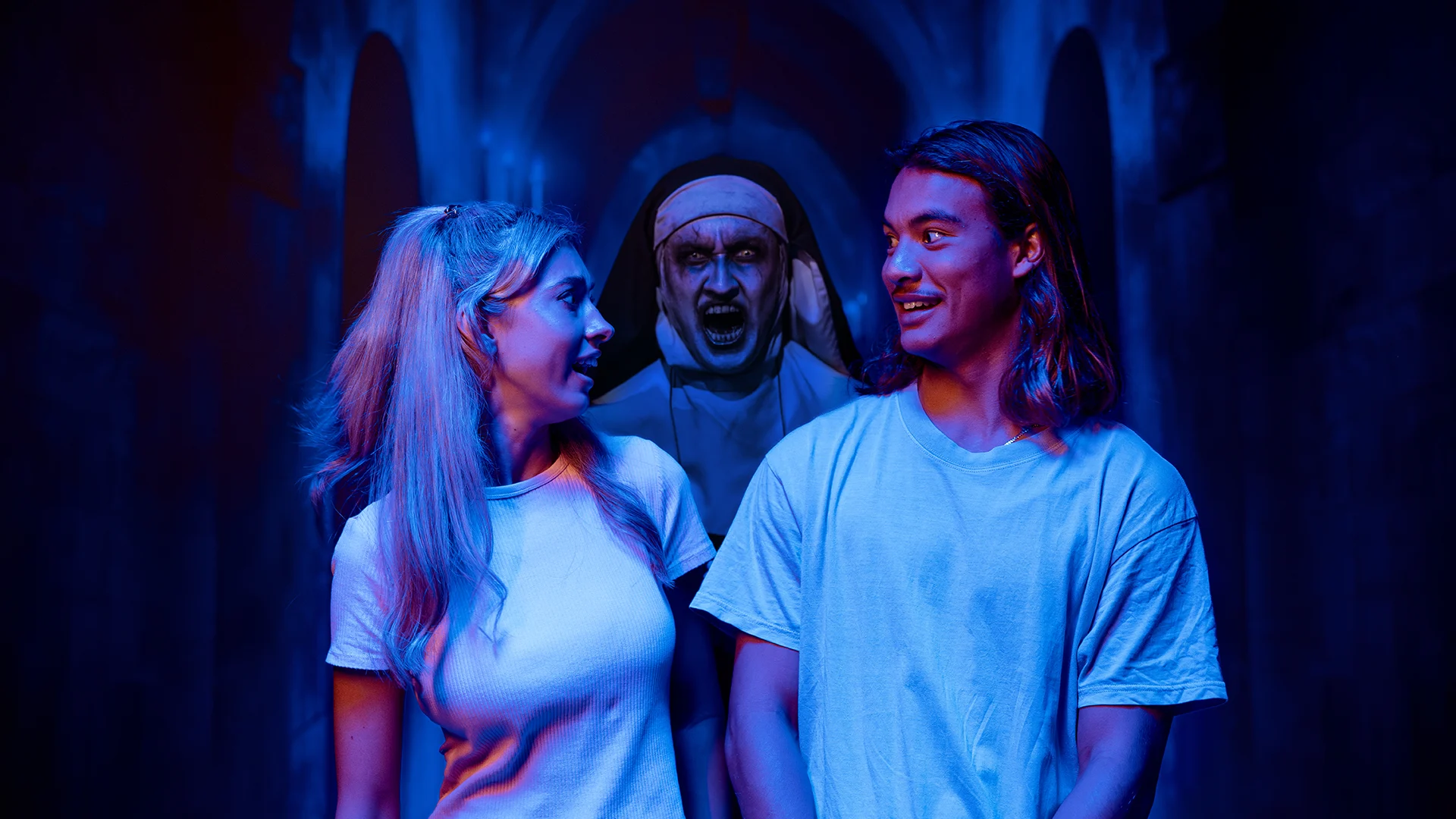 A young man and woman in white shirts smile at each other, unaware of a frightening, ghostly nun looming behind them in a dark, eerie hallway with blue lighting.