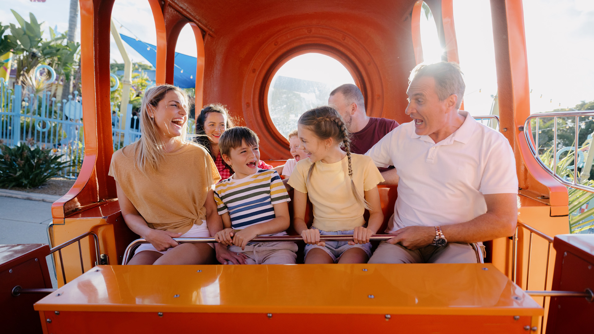 A group of adults and children sit together inside a bright orange amusement park ride, smiling and laughing.