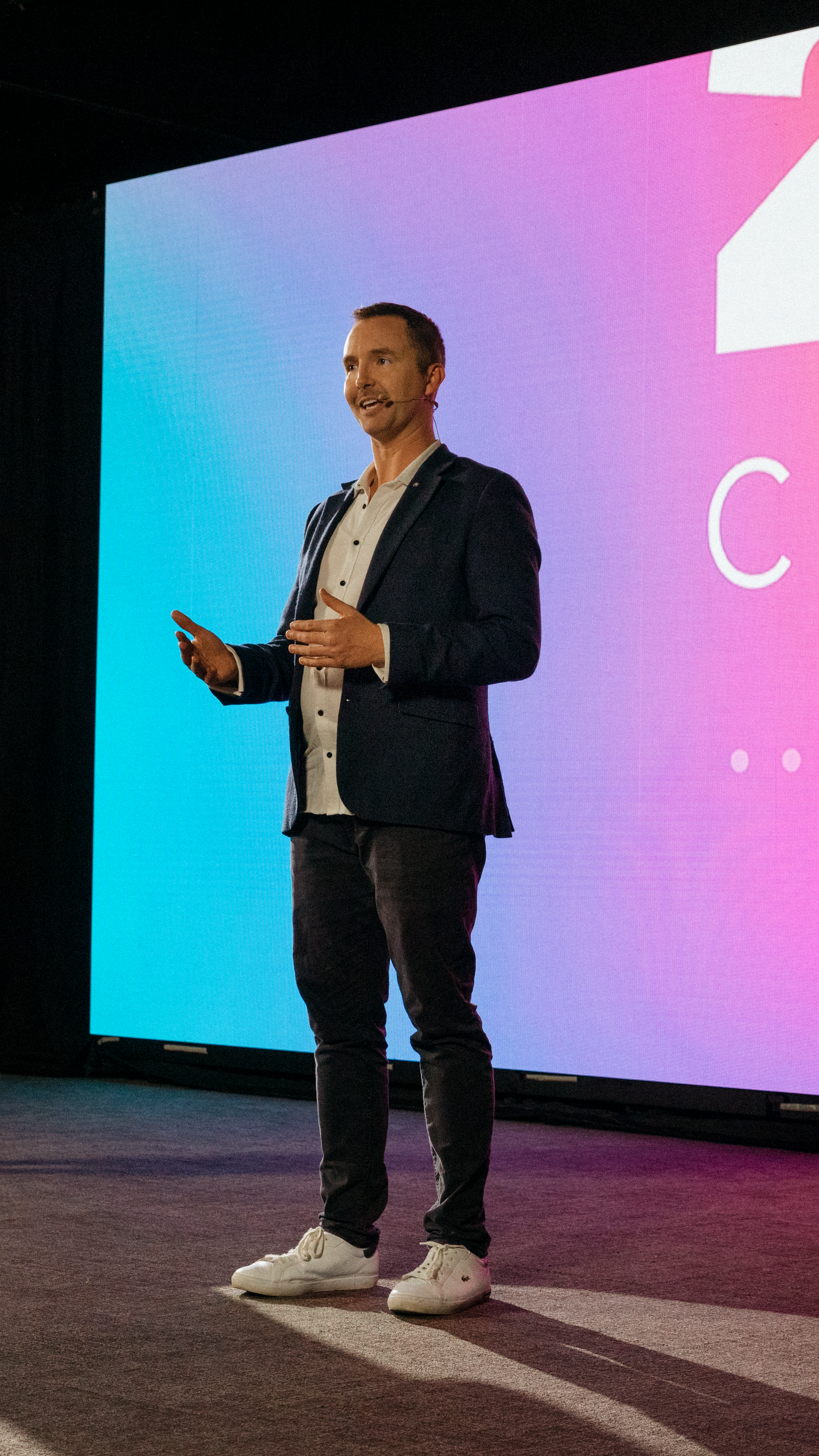 Man in casual attire speaking on stage with a colorful background screen.
