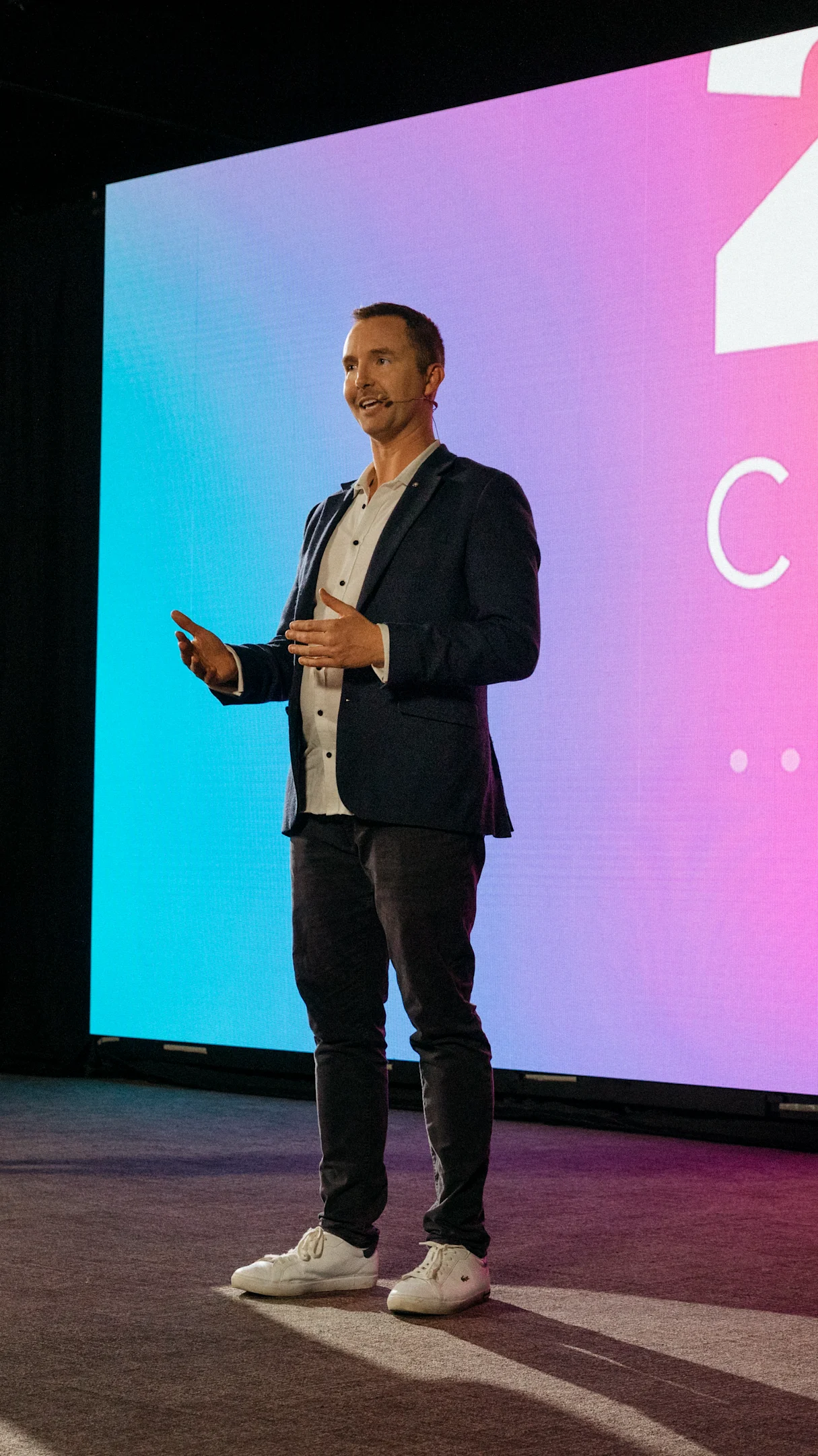 Man in casual attire speaking on stage with a colorful background screen.