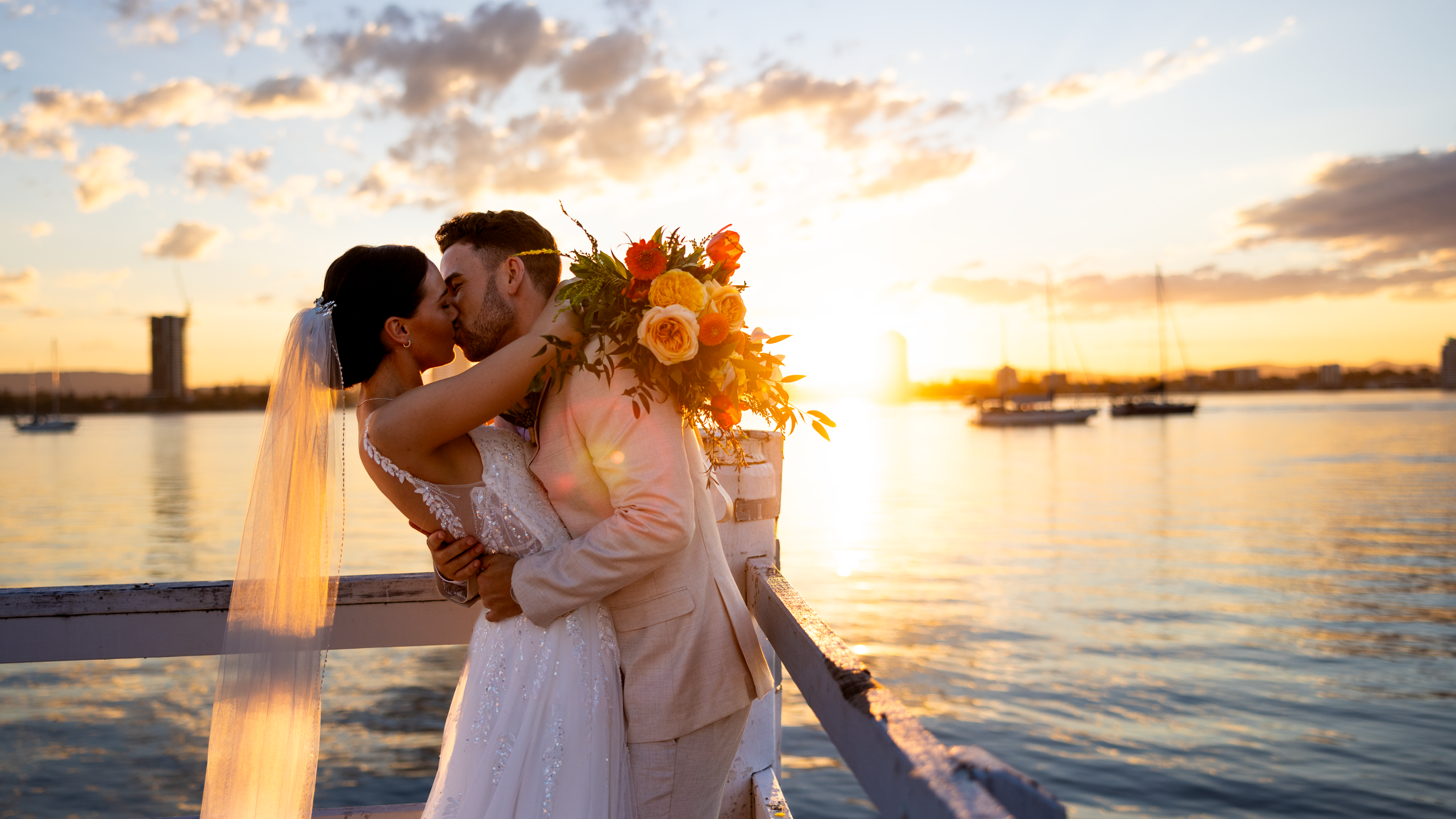 A couple in wedding attire kisses on a pier during sunset, with a bouquet in hand and sailboats on the water in the background.
