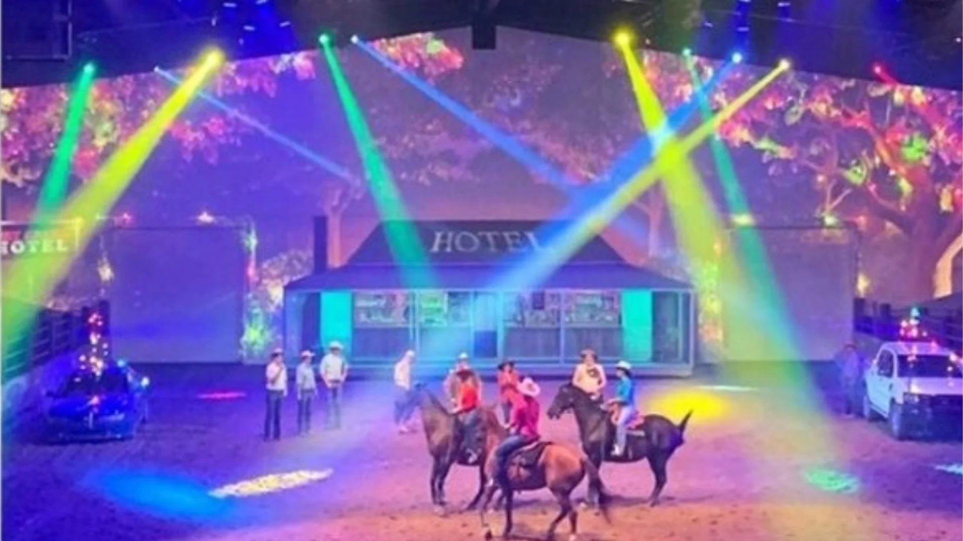 Performers on horseback and standing on stage are illuminated by colorful spotlights in front of a building labeled "HOTEL," with audience seating and two white vehicles on either side.