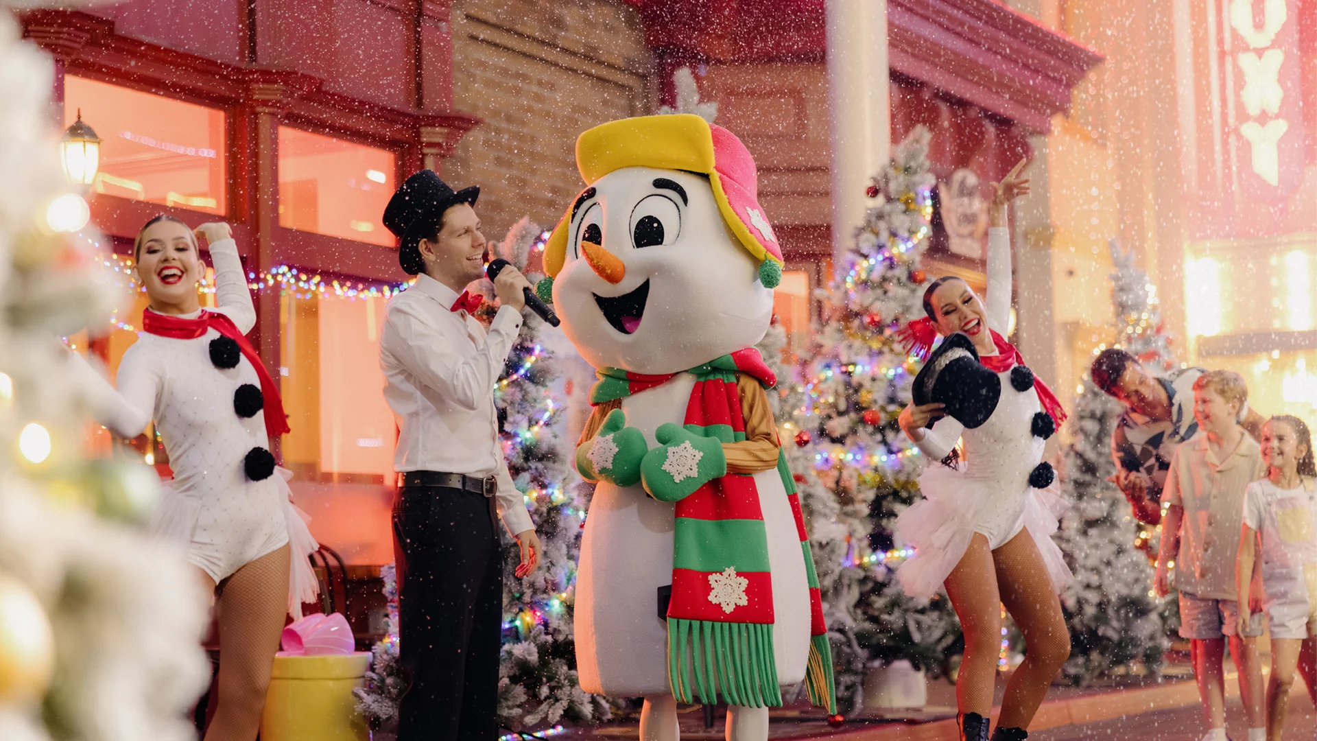 People in festive costumes, including a large snowman mascot, dance and celebrate in a snowy, Christmas-themed outdoor setting with decorated trees and bright lights in the background.