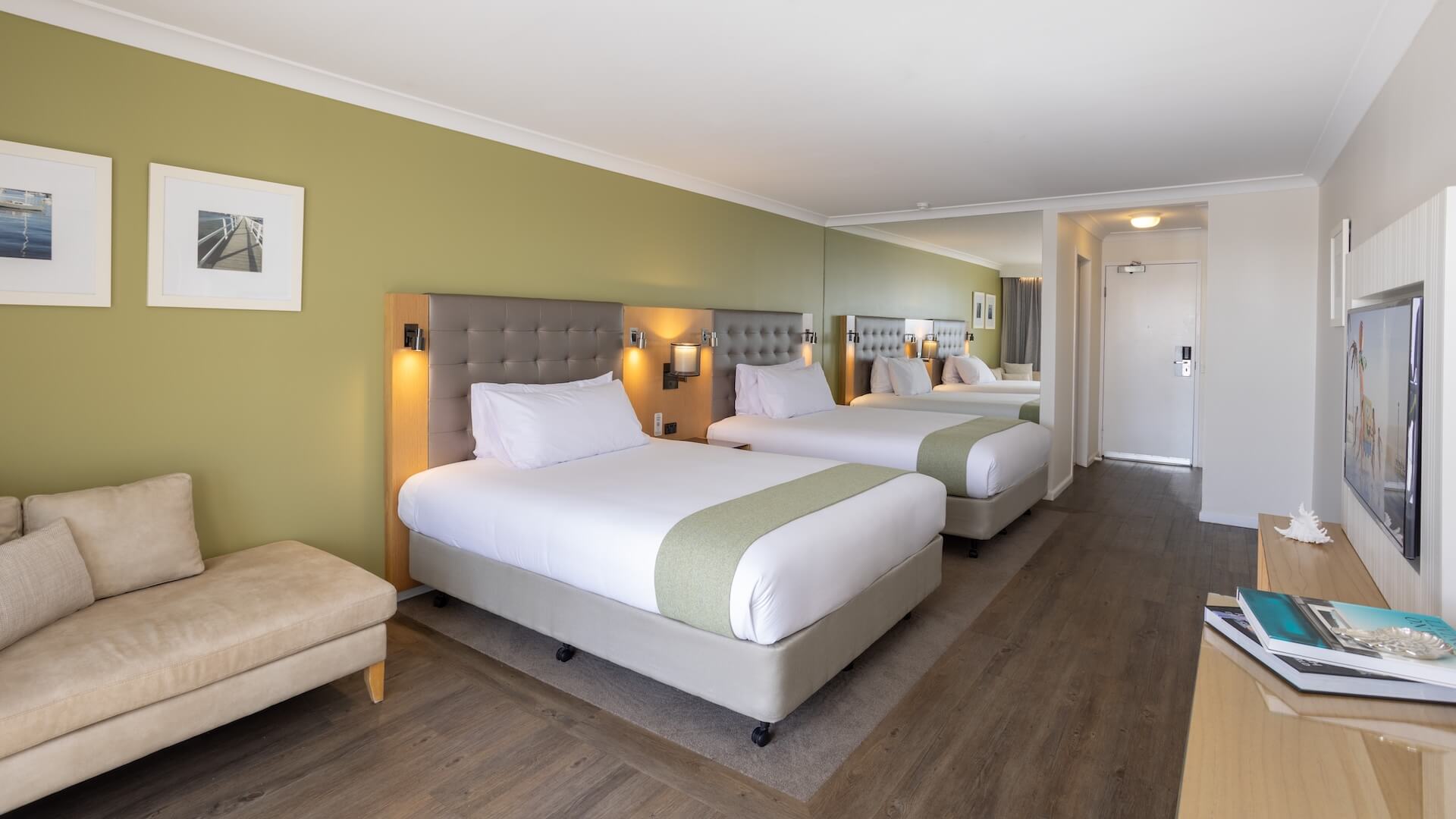 Hotel room with two double beds, a green accent wall, sofa, wooden floor, white bedding, mirror, framed pictures, and a hallway leading to a door.