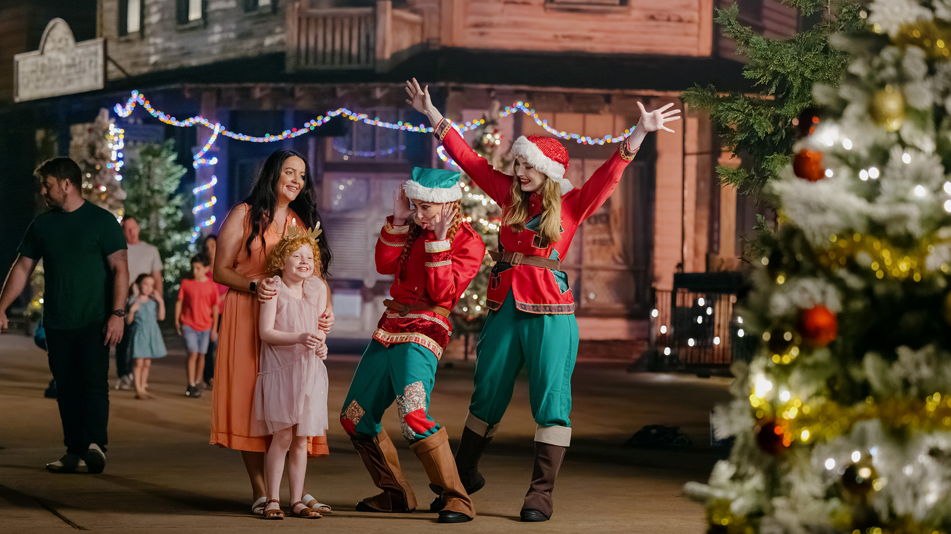 A family with a young girl smiles next to two people dressed as elves in festive costumes. They all pose joyfully near a decorated Christmas tree, with holiday lights and wooden buildings in the background.