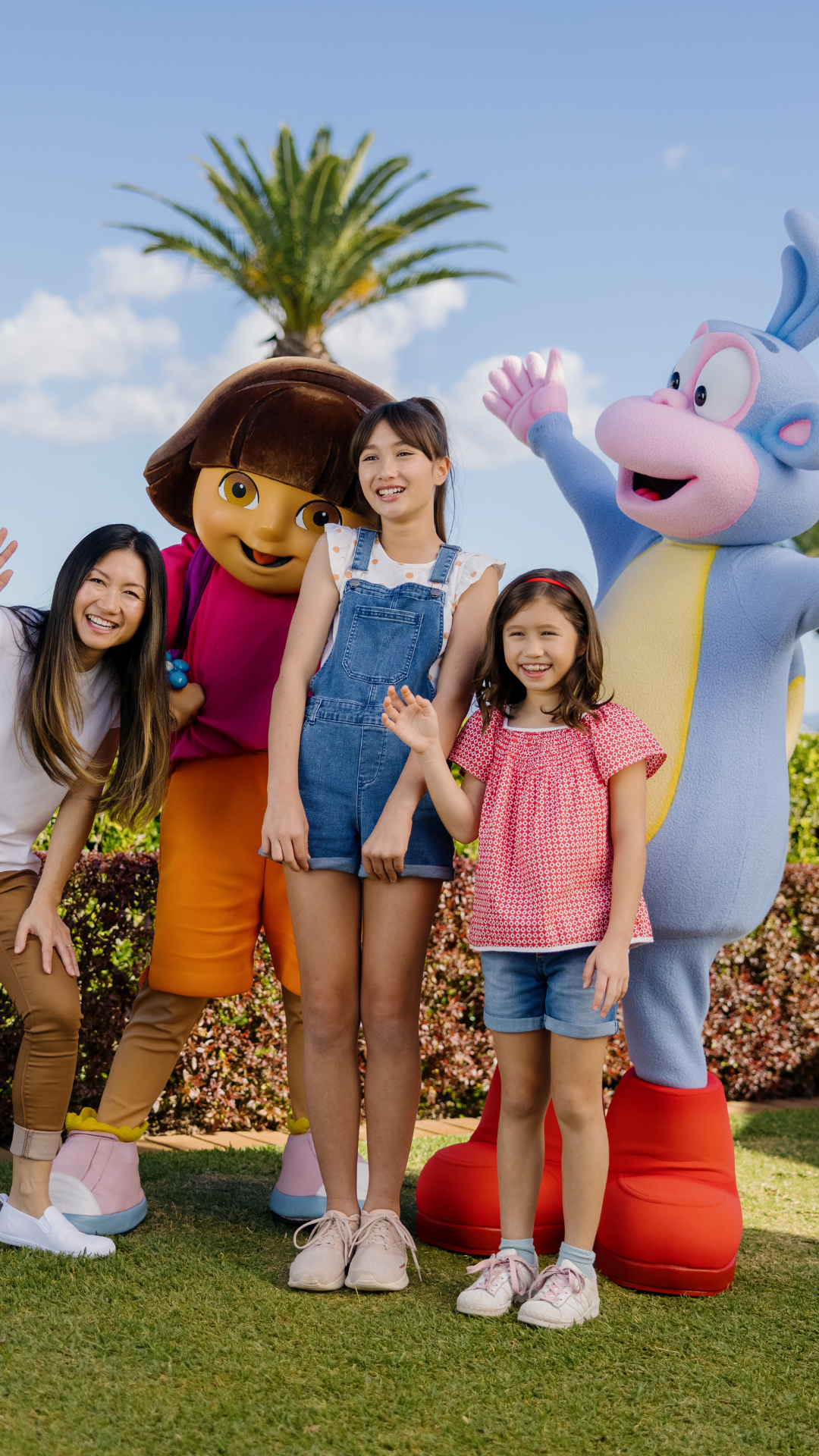 Two children and a woman pose outdoors with people dressed as Dora the Explorer and Boots the Monkey, smiling under a sunny sky with palm trees in the background.