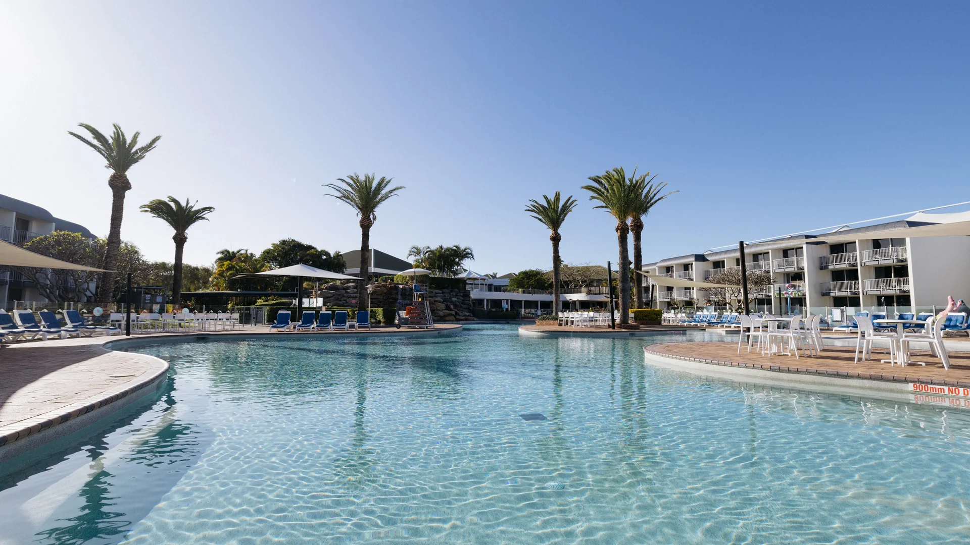 A clear outdoor swimming pool surrounded by lounge chairs, umbrellas, and palm trees, with resort buildings in the background under a bright blue sky.