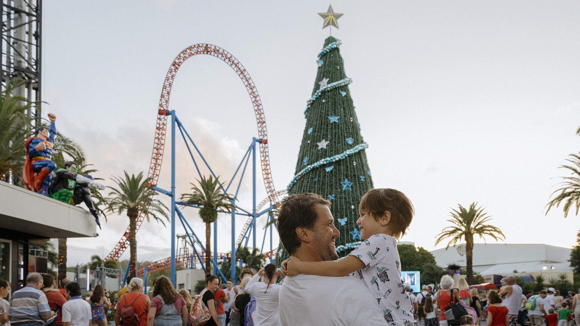 A man holds a young child while standing in a crowded amusement park decorated for Christmas, featuring a large Christmas tree, a roller coaster, palm trees, and festive atmosphere. 
