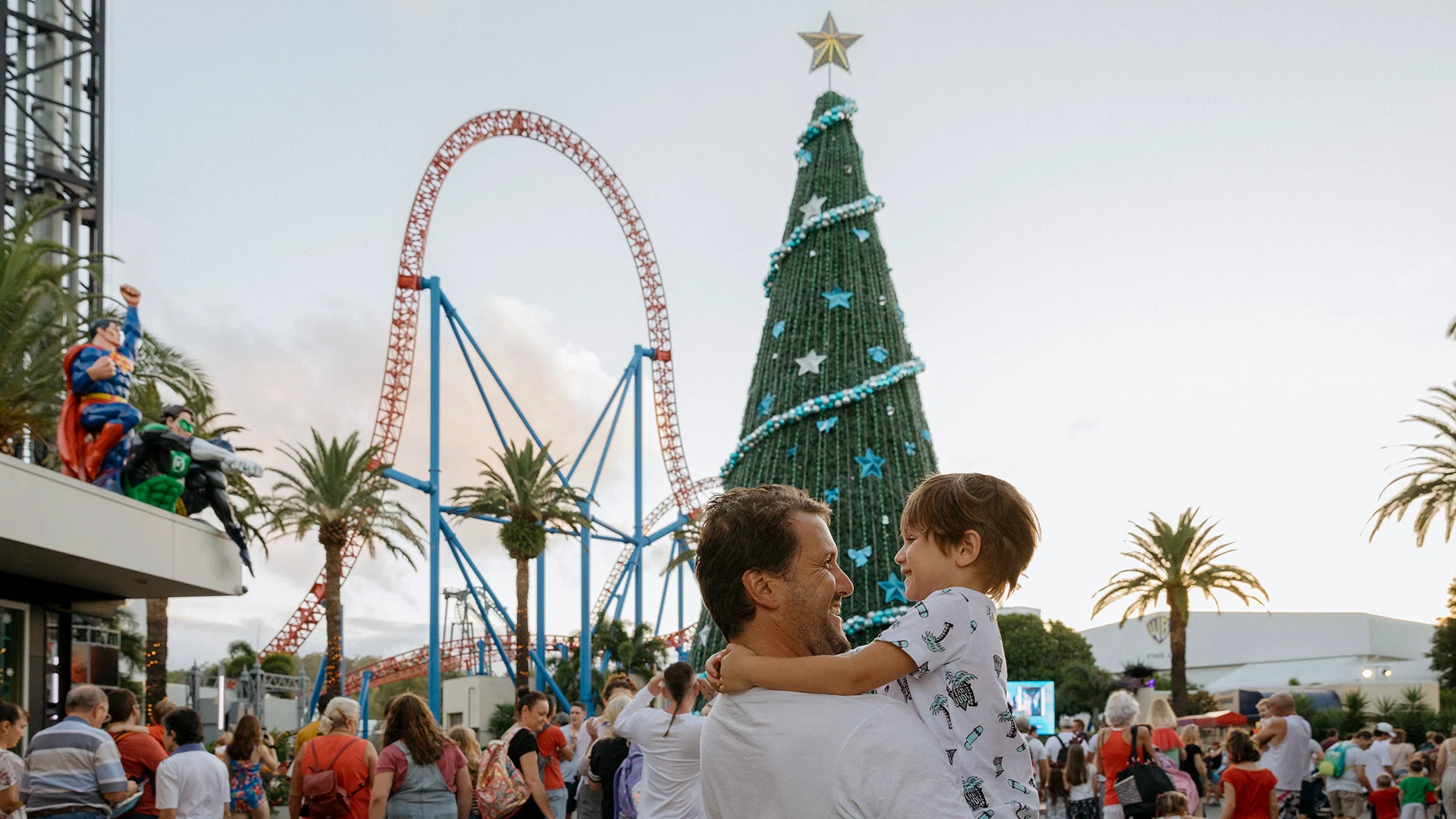 A man holds a young child while standing in a crowded amusement park decorated for Christmas, featuring a large Christmas tree, a roller coaster, palm trees, and festive atmosphere.