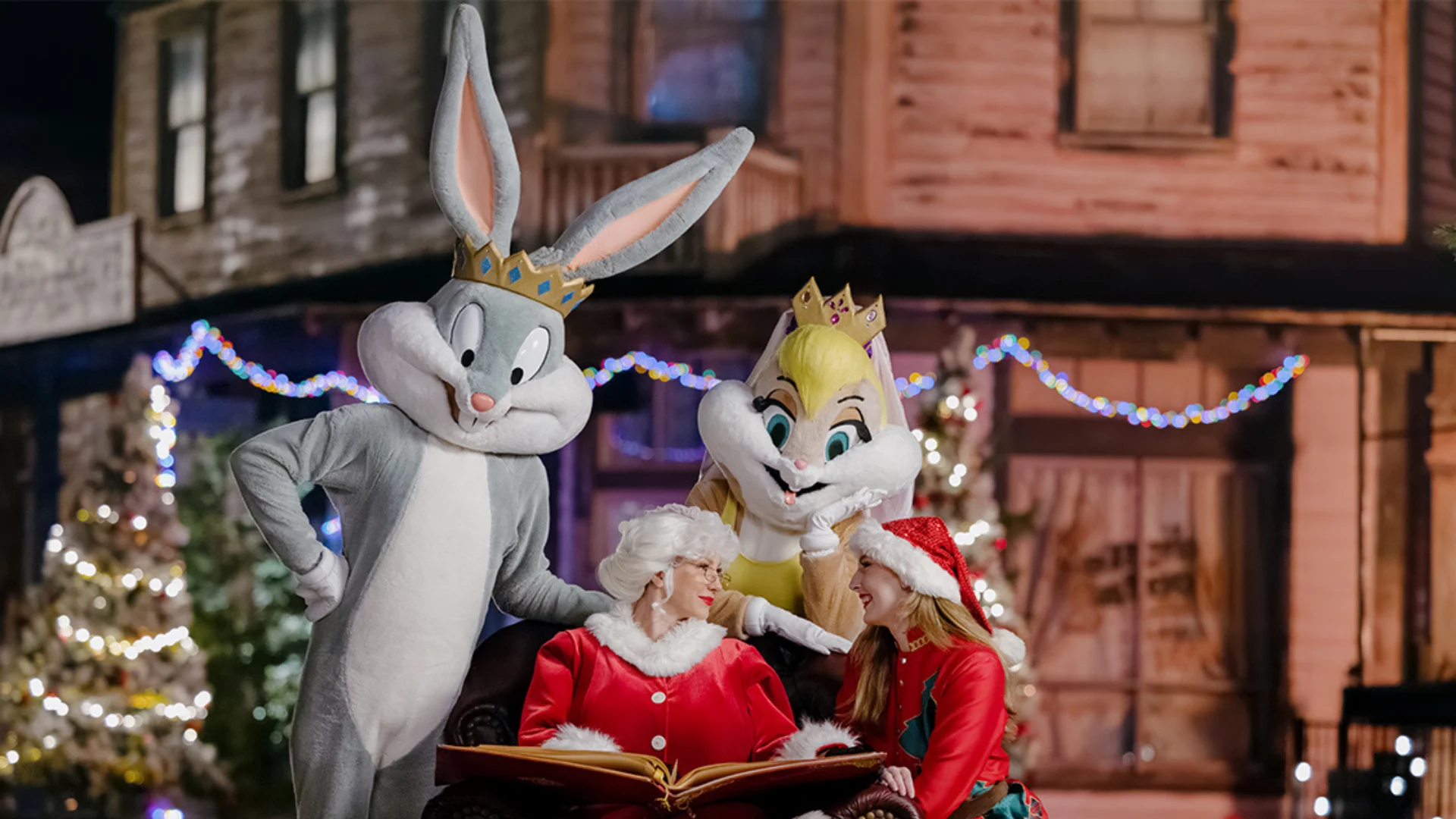 Bugs Bunny and Lola Bunny, both wearing crowns, stand beside two women dressed in Santa outfits, who are sitting with an open book. Christmas lights and decorated trees are in the background.