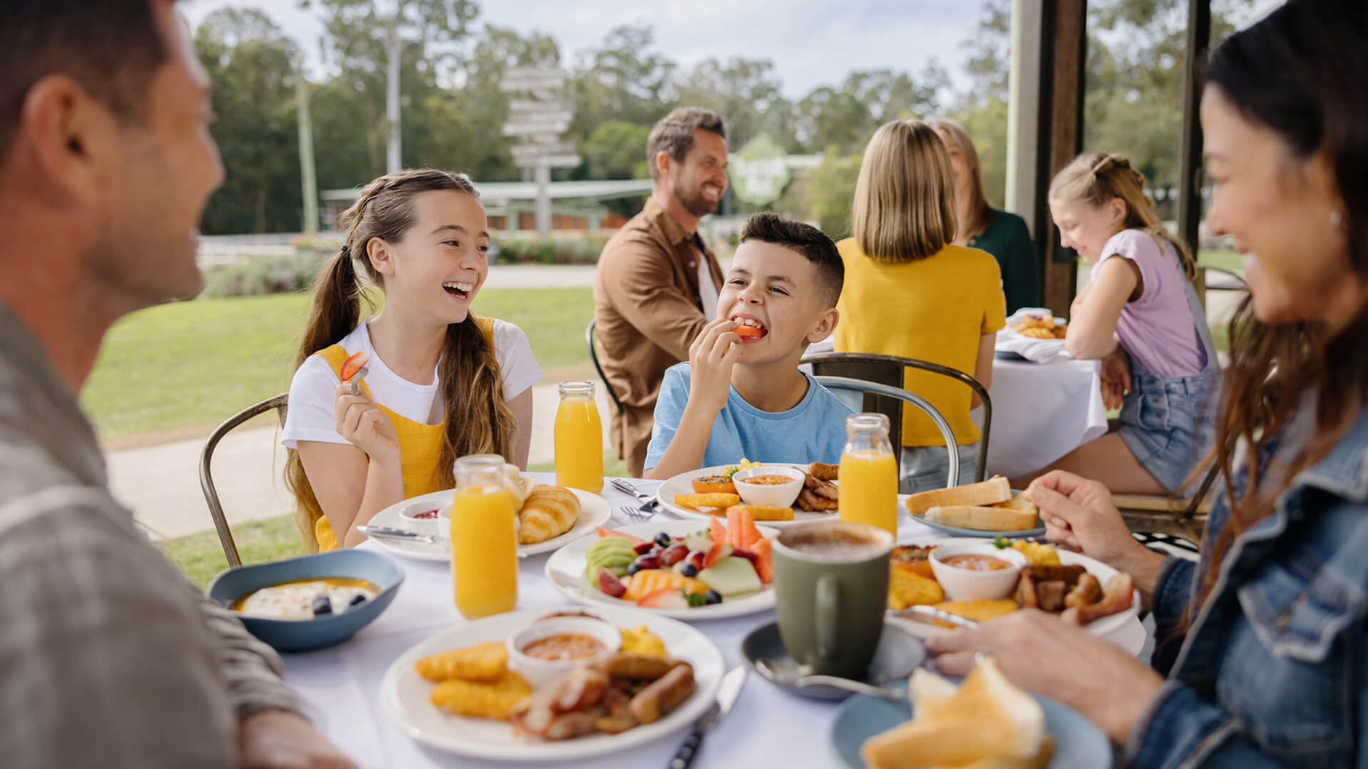 A family sits at an outdoor table enjoying breakfast foods and orange juice, laughing and talking together on a sunny day.